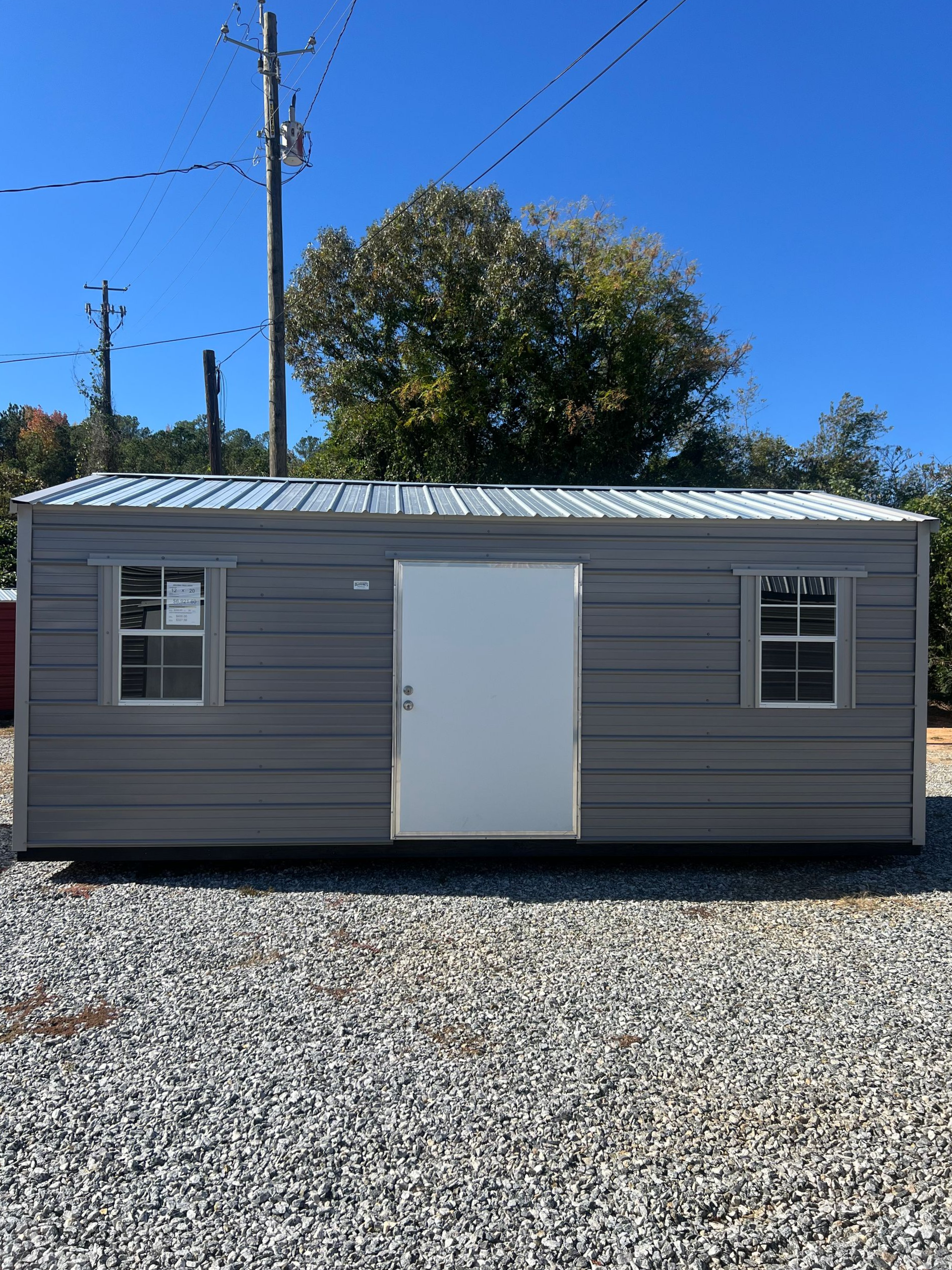 Gray metal shed with a white door and two windows on a gravel lot under a blue sky.