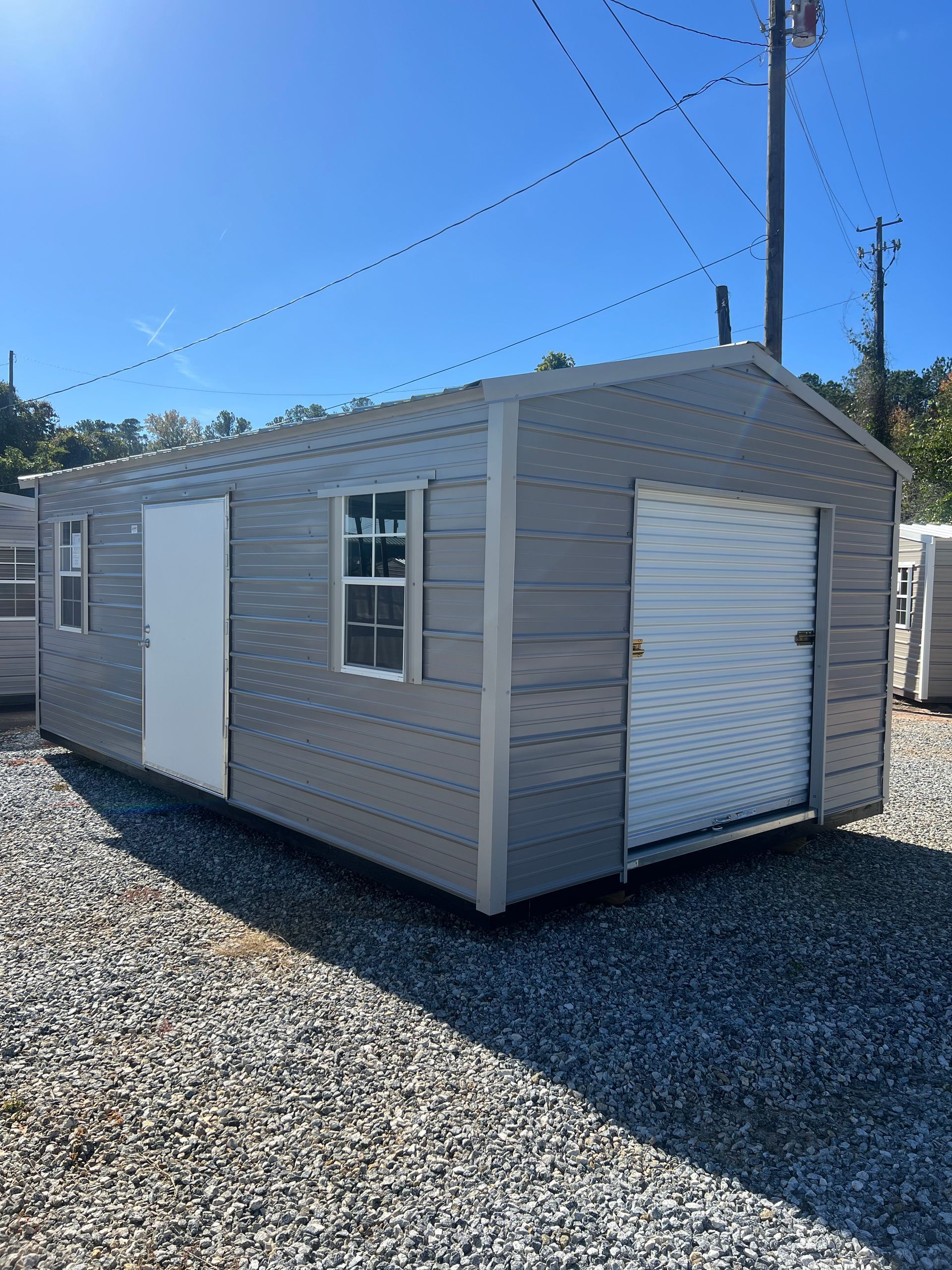 Gray metal shed with a roll-up garage door, a side door, and two windows on a gravel lot under a blue sky.