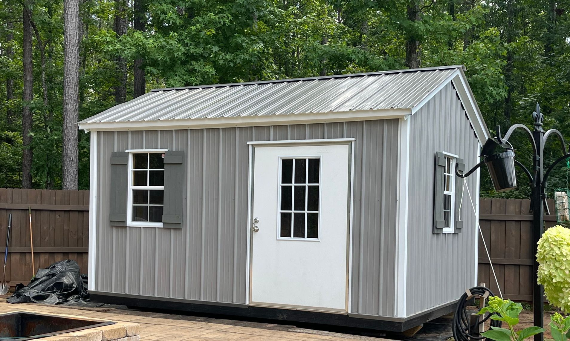Gray shed with a white door and shutters; metal roof against a backdrop of trees.