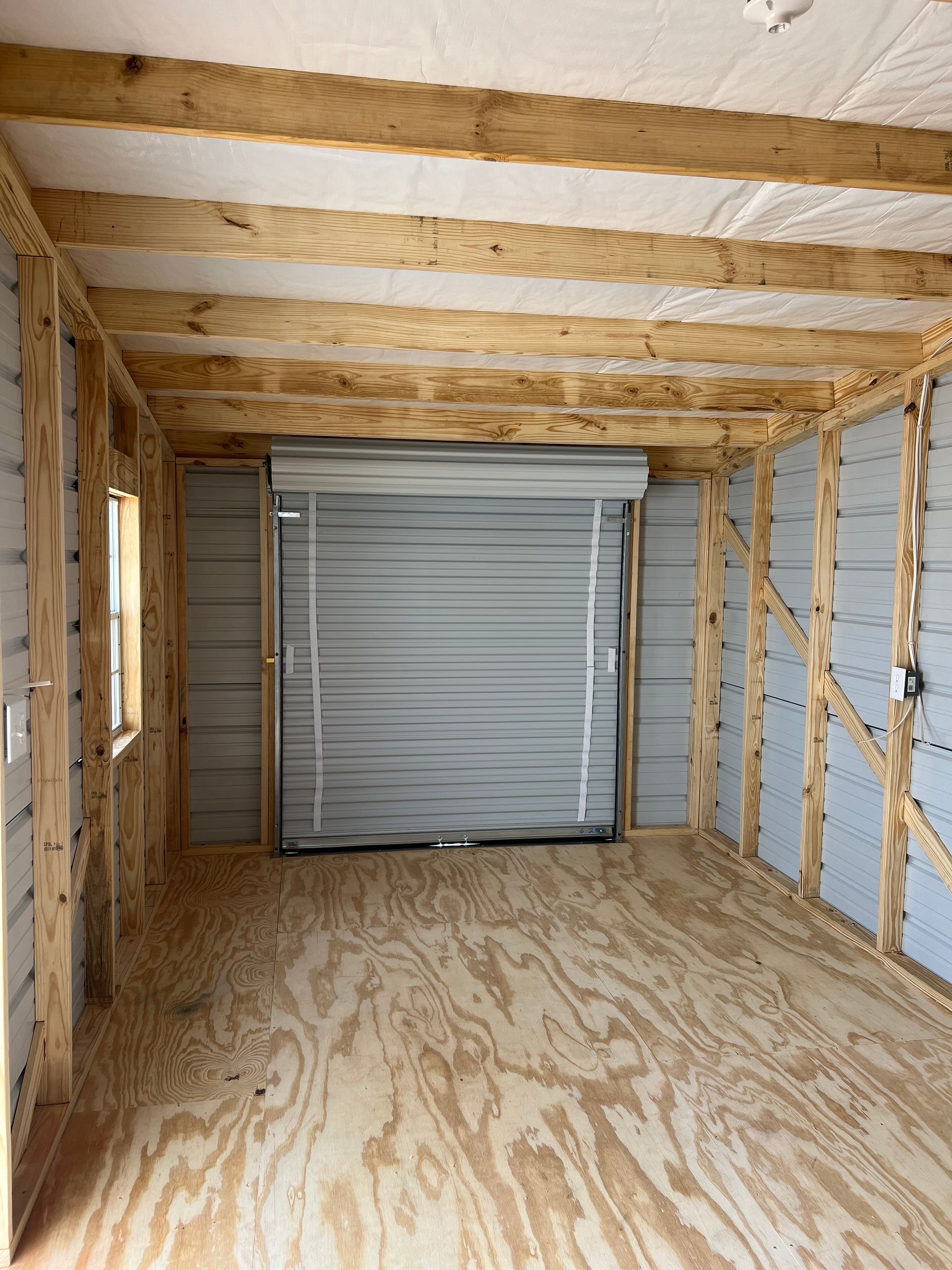 Interior of a shed with a roll-up door, window, and wood beams.  Gray metal walls and plywood floor.