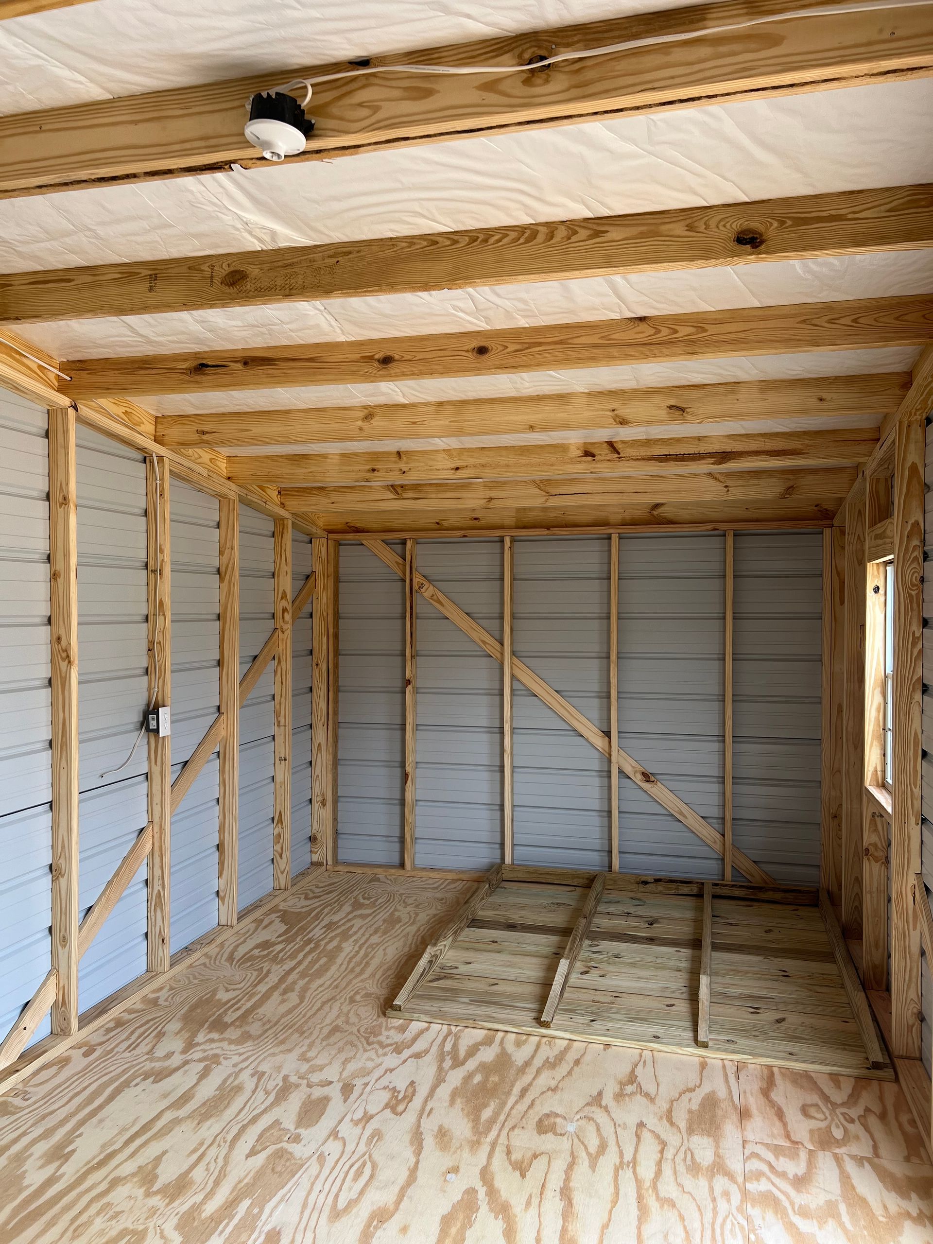 Interior of a wooden structure with exposed beams, insulated walls, and plywood floor. A light fixture is mounted on the ceiling.
