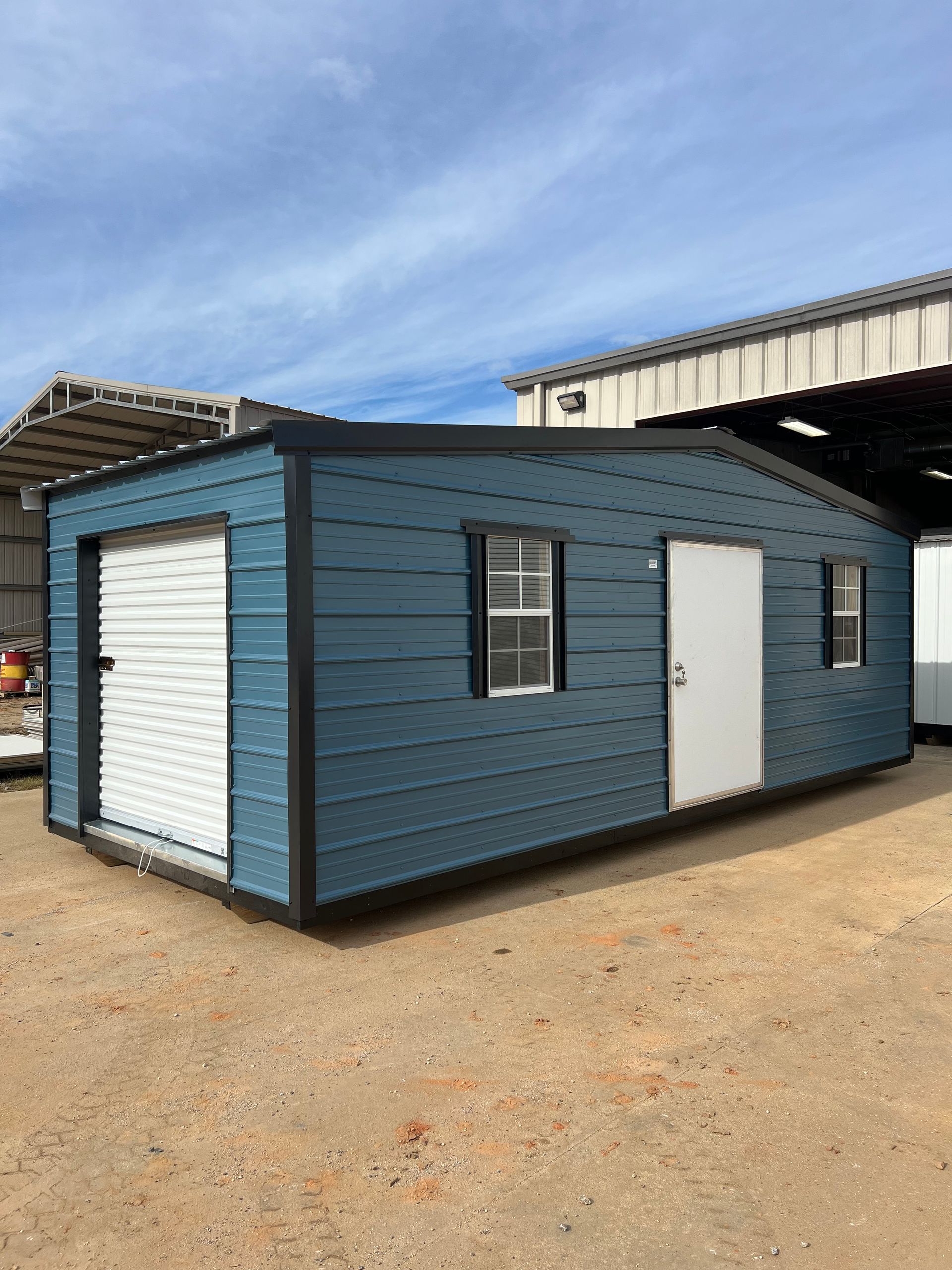 Blue metal shed with garage door, white door, and windows. Black trim, sunny outdoor setting.