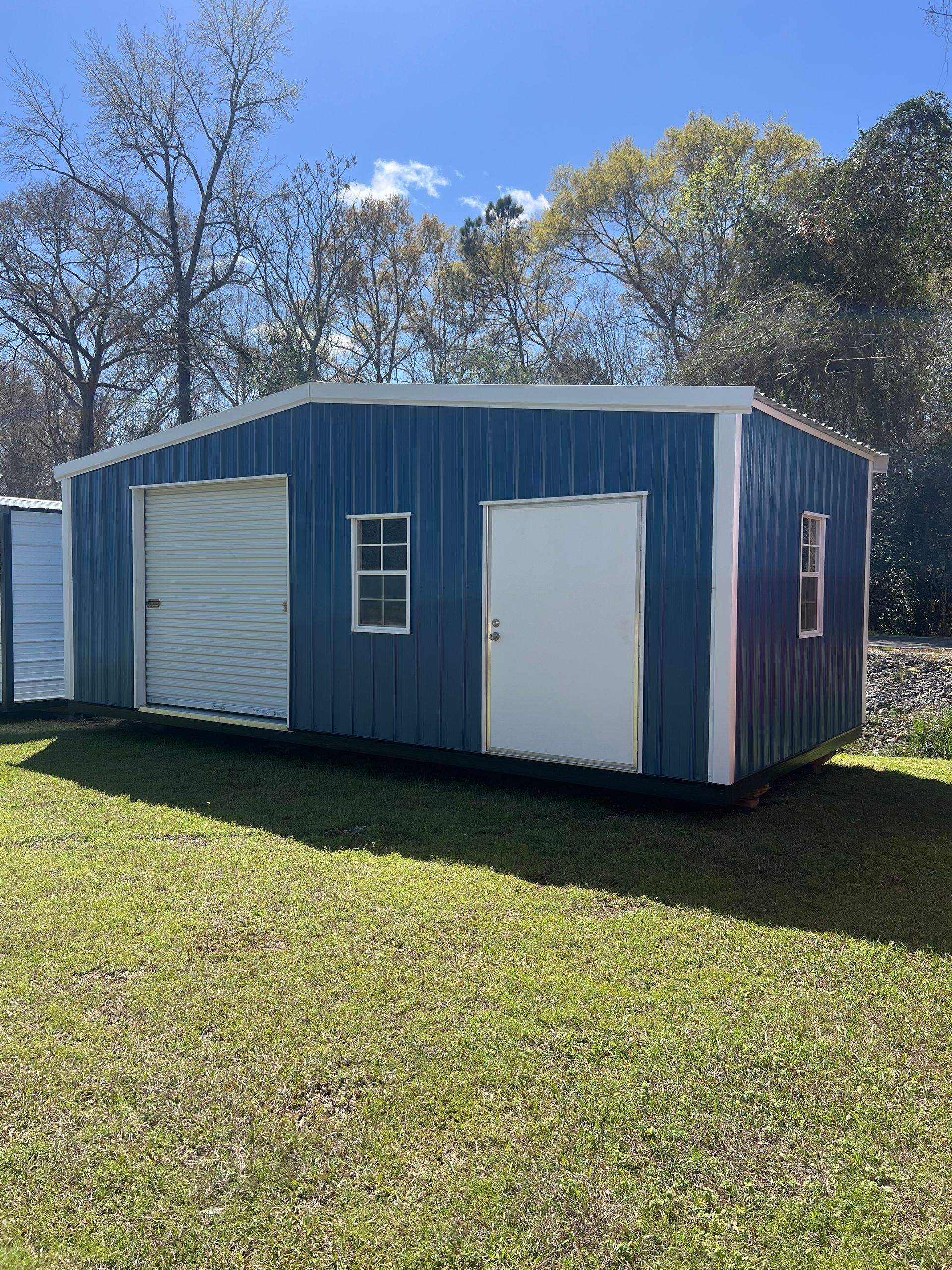 A blue metal utility shed with a white roll-up door, a single door, and two windows, situated in a grassy yard.