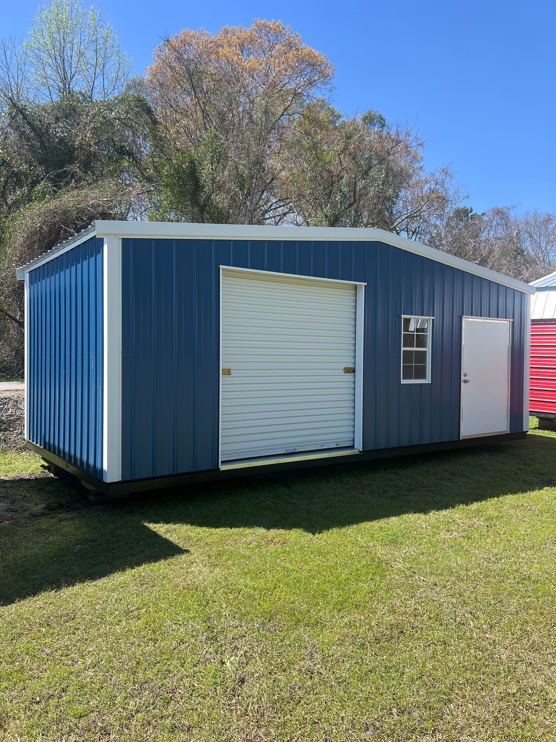 A blue metal storage shed with a white roll-up door, a window, and a white entry door, sitting on a grassy lawn.