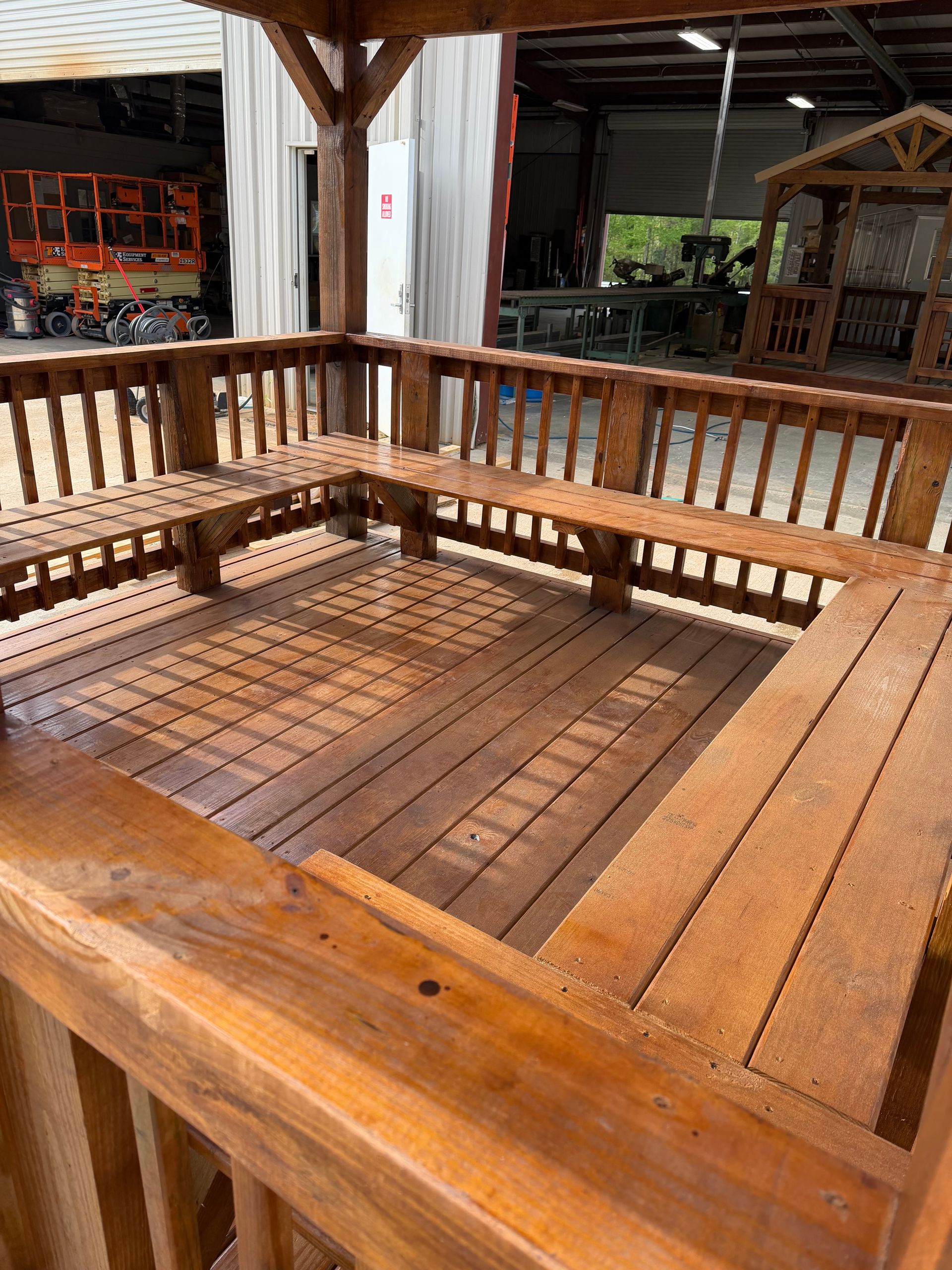 Wooden gazebo interior featuring built-in bench seating along the perimeter and a planked deck floor, in a workshop.