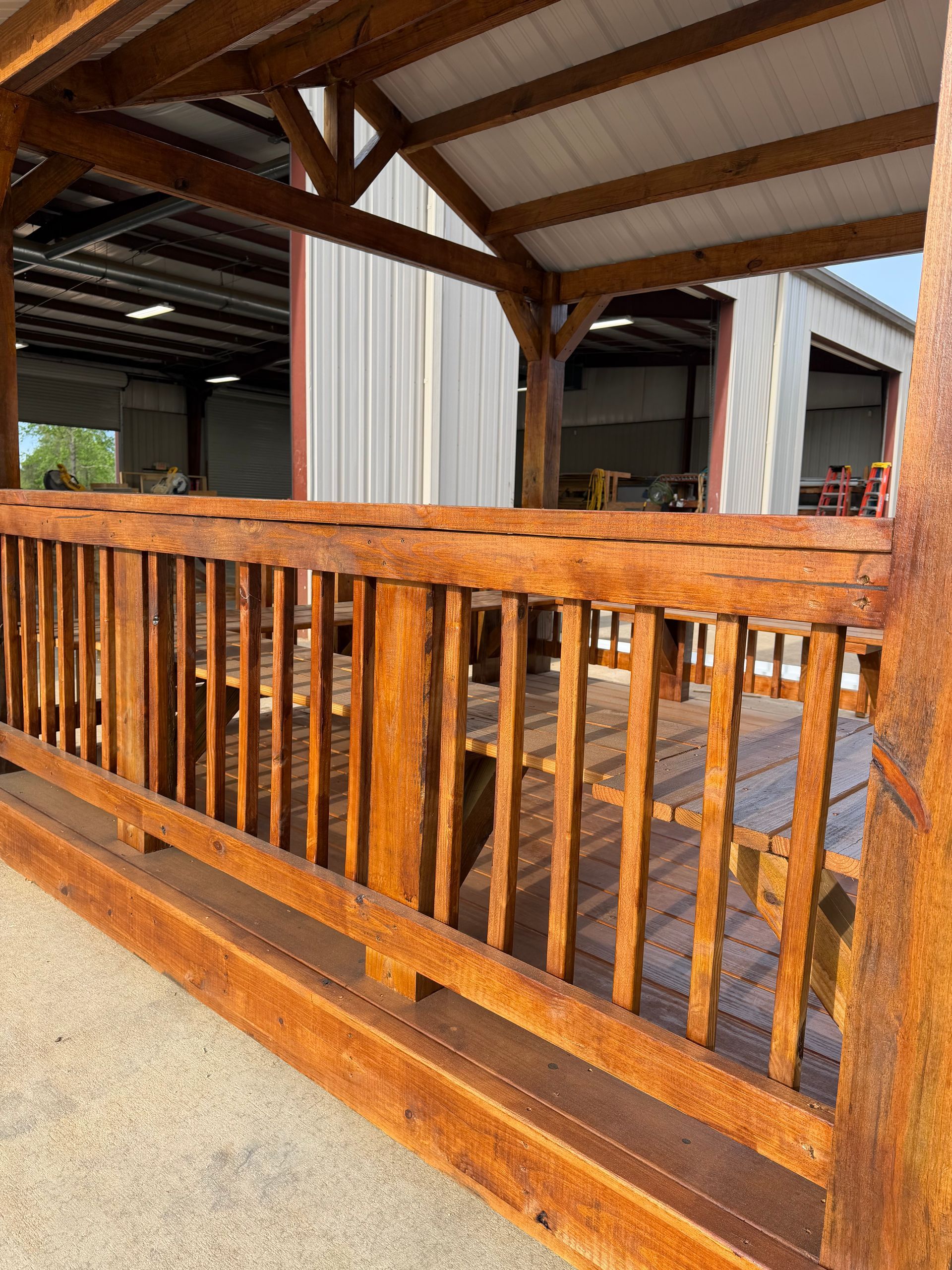 Stained wooden pavilion with a peaked roof, featuring a railed platform and picnic-style seating, set in an open warehouse.