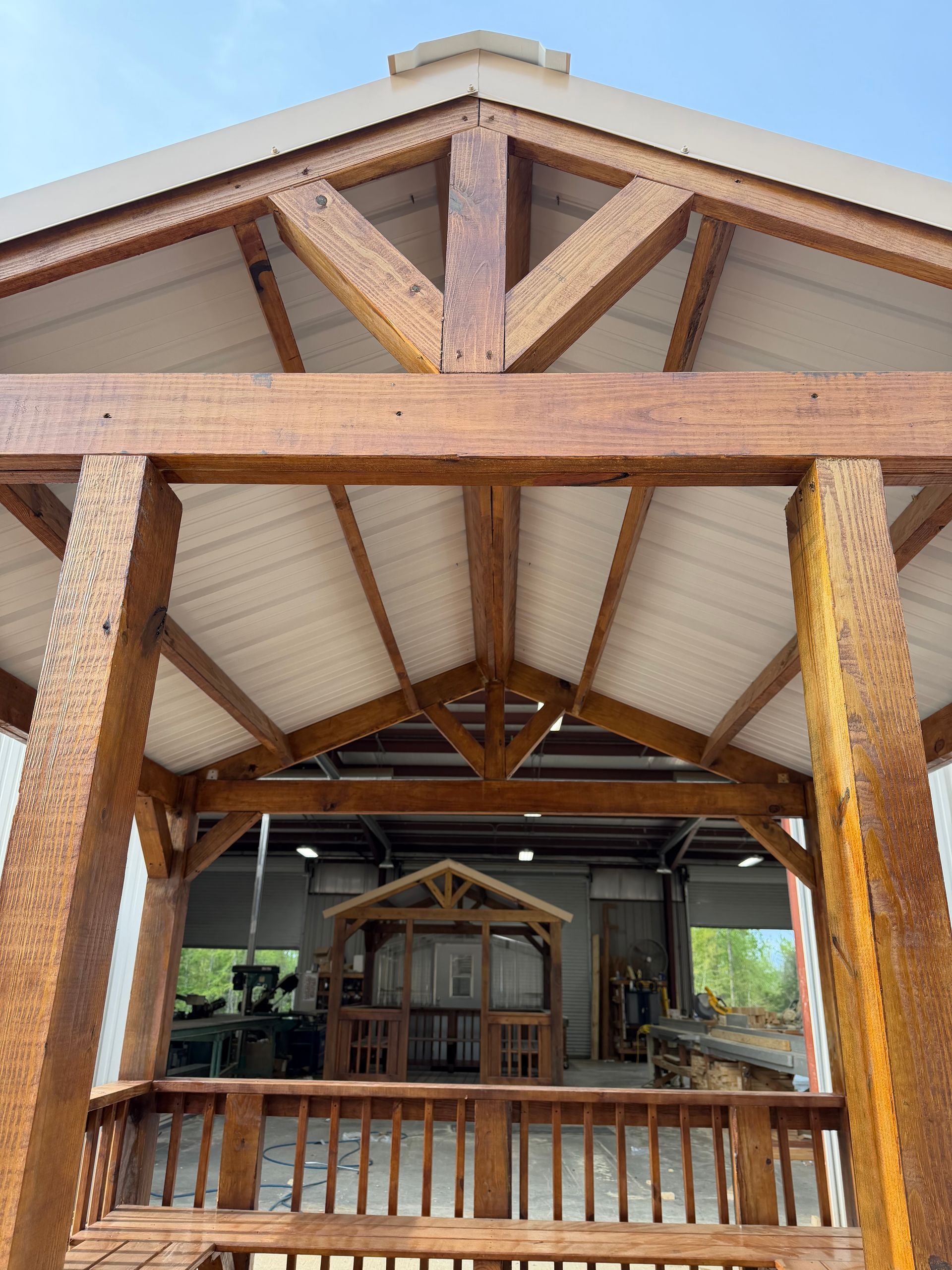 A wooden gazebo frame with a peaked roof stands in a workshop, showcasing timber beams and a metal ceiling.