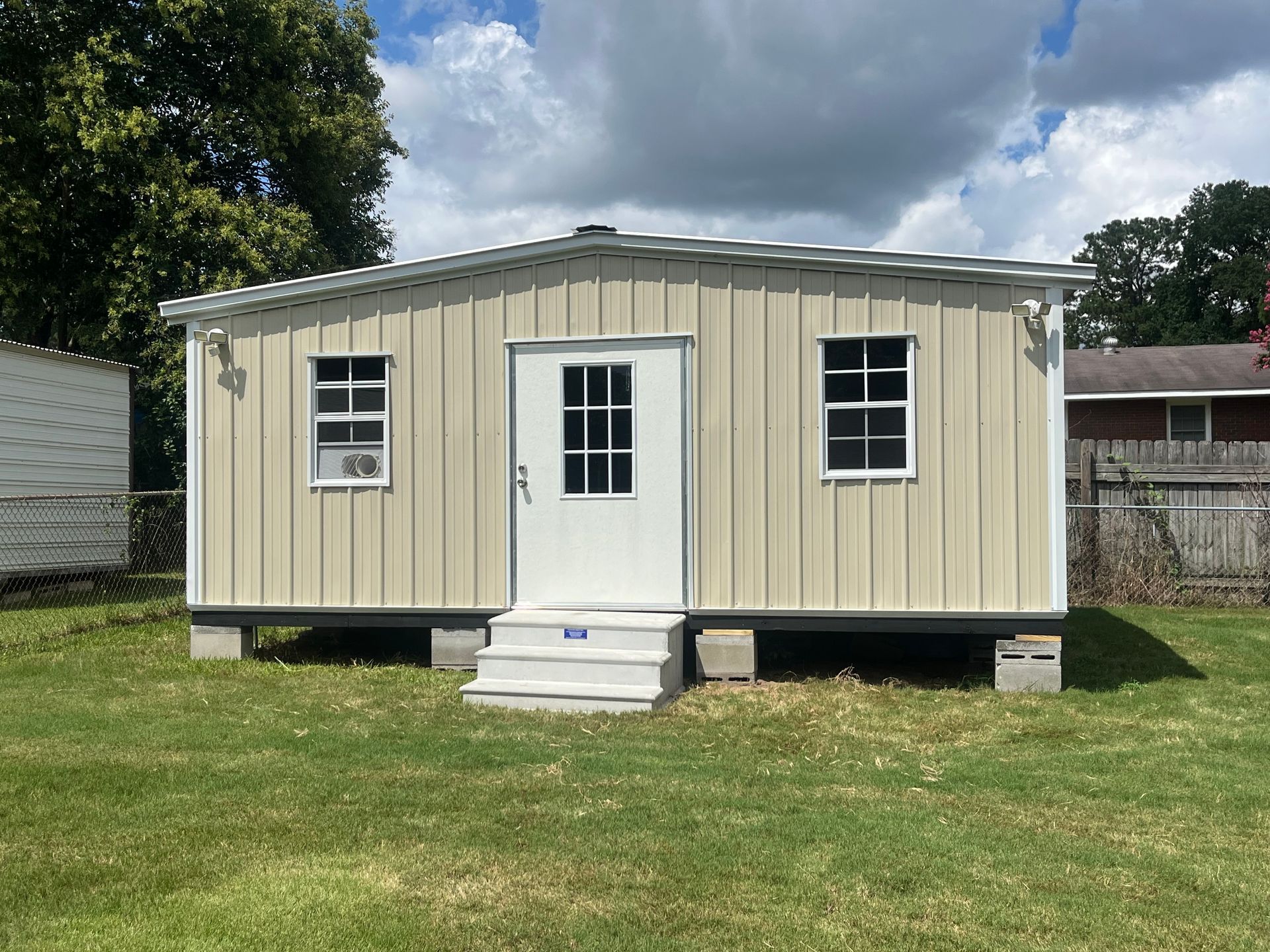 Tan metal shed with white door and windows on a grassy lawn under a cloudy sky.