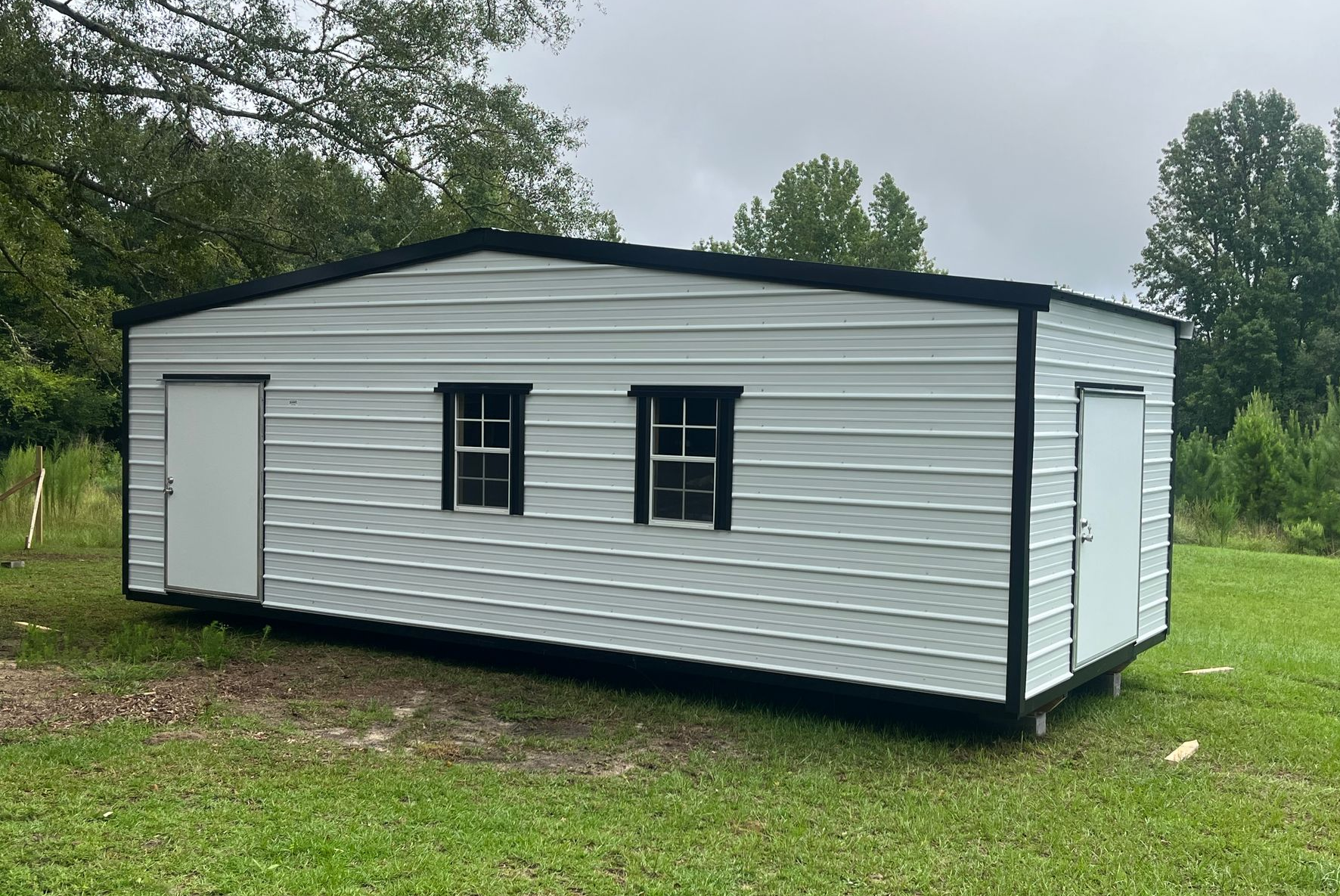 White metal building with black trim, two windows, and two doors, set in a grassy area with trees.