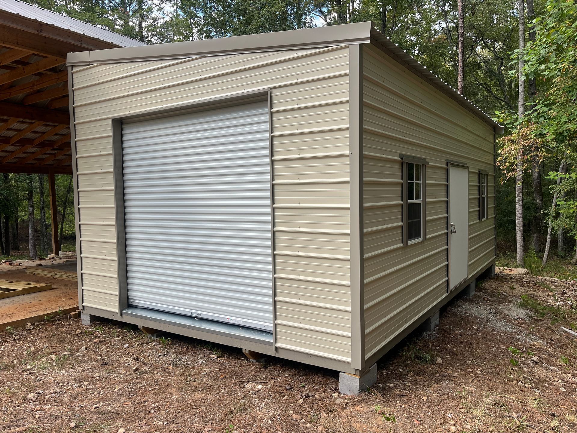 Tan metal shed with a roll-up door, windows, and a side door, set in a wooded area.