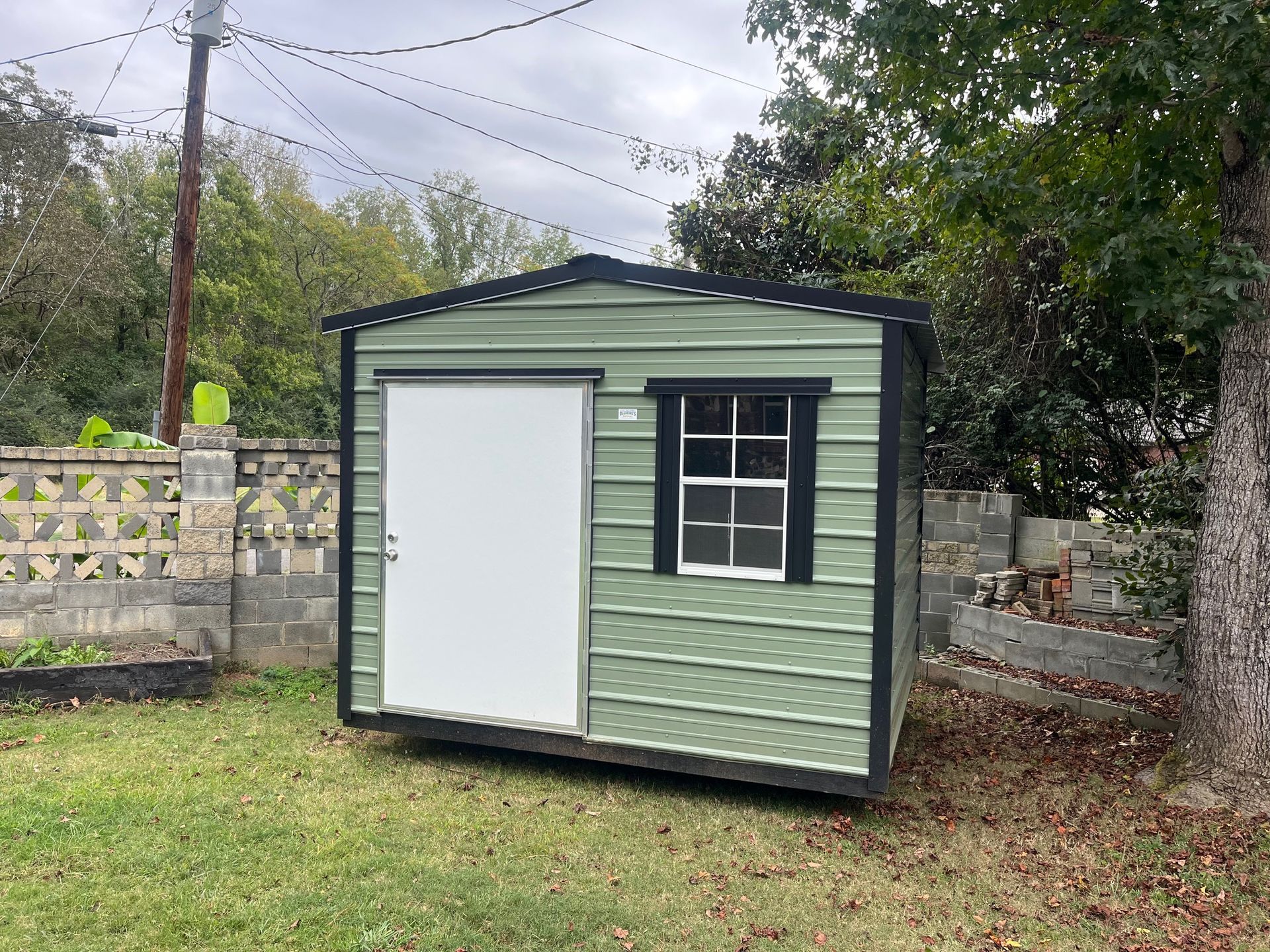 Green storage shed with black trim, a white door, and a window in a grassy yard.