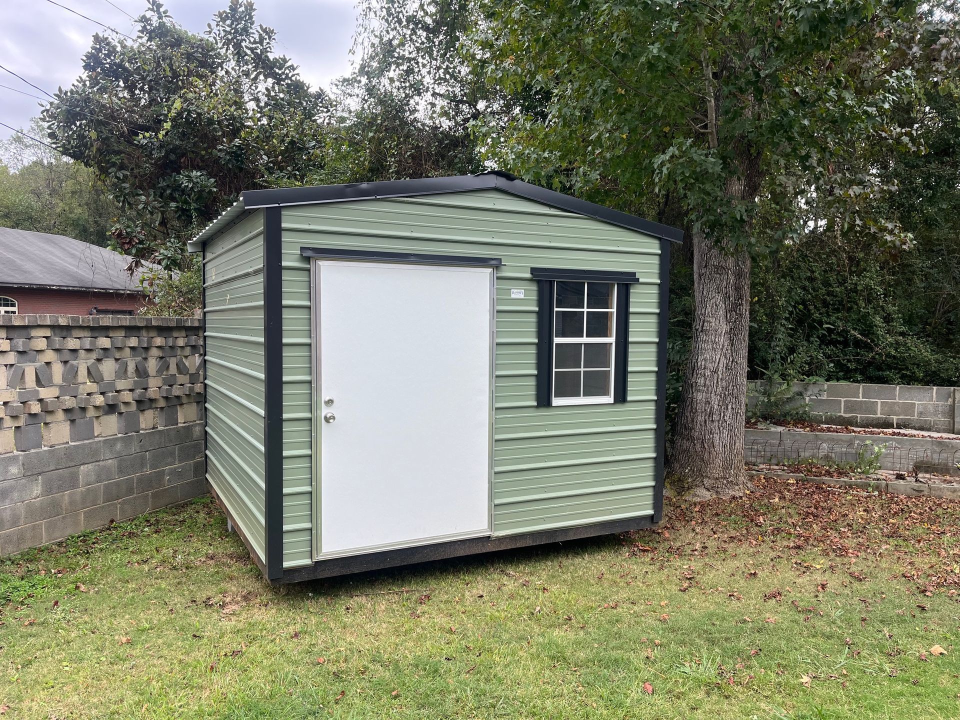 Green storage shed with white door and window, set on grass near a tree.
