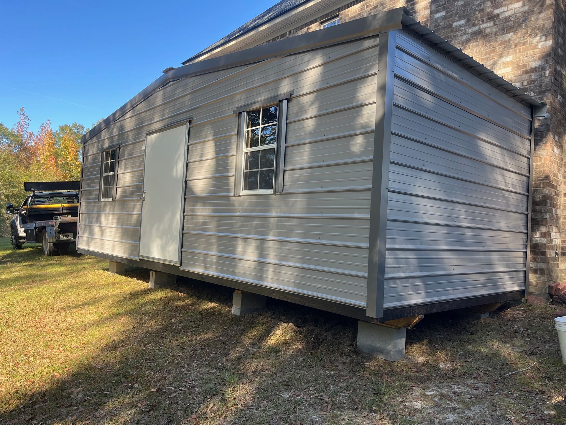 Gray metal shed with a door and windows, set on concrete blocks, outdoors on grass.
