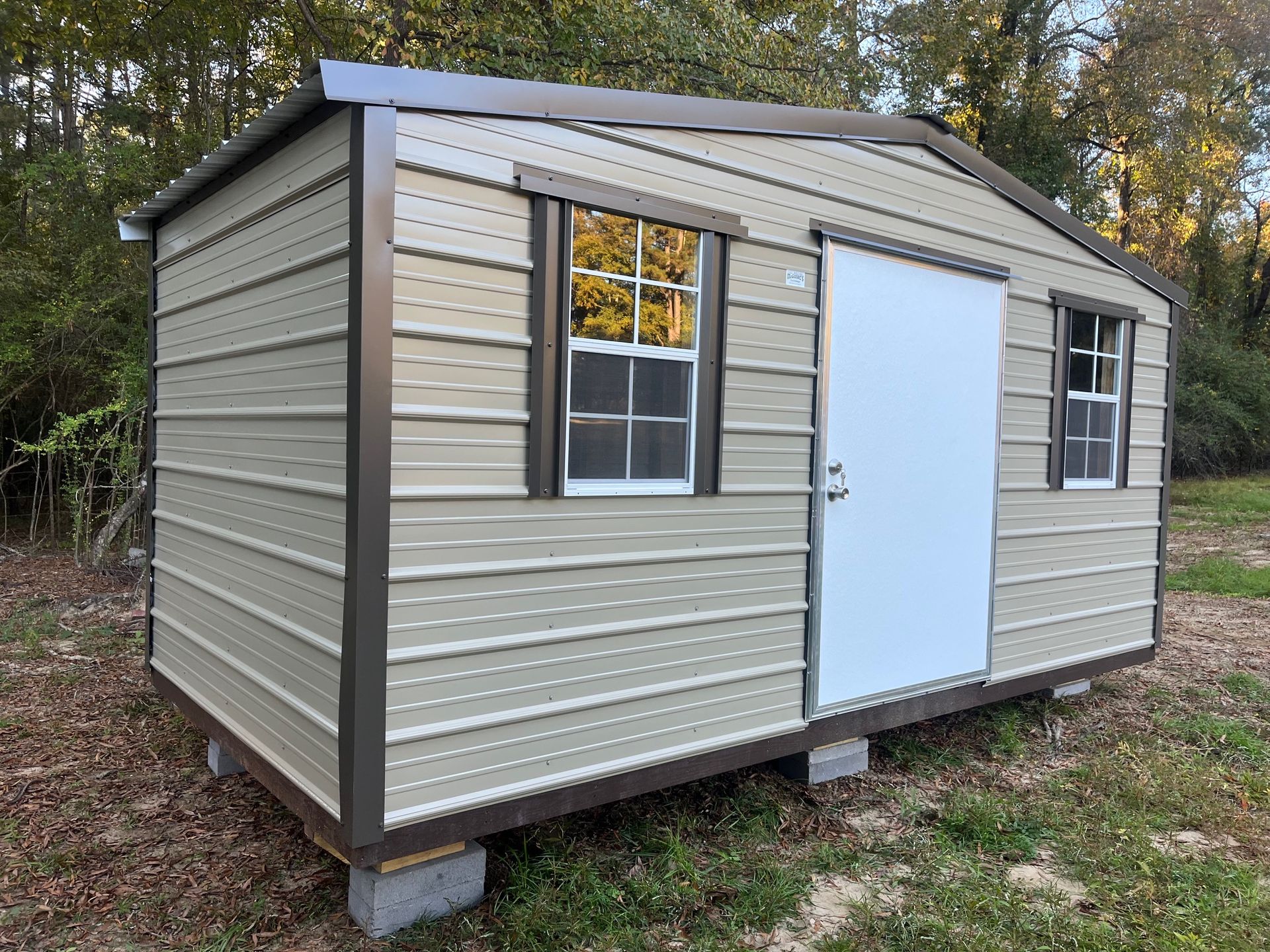 Tan metal storage shed with brown trim, door, and windows, set on concrete blocks outdoors.
