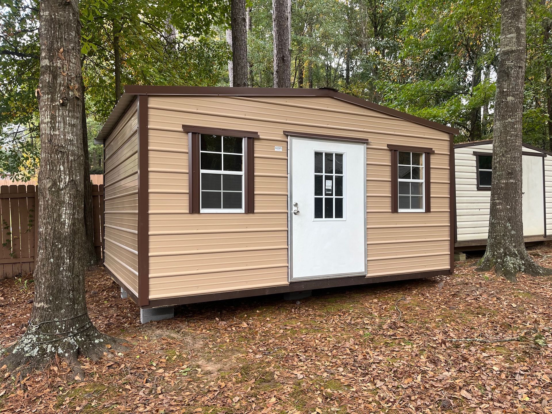 Tan shed with brown trim, white door, and windows in a wooded area.