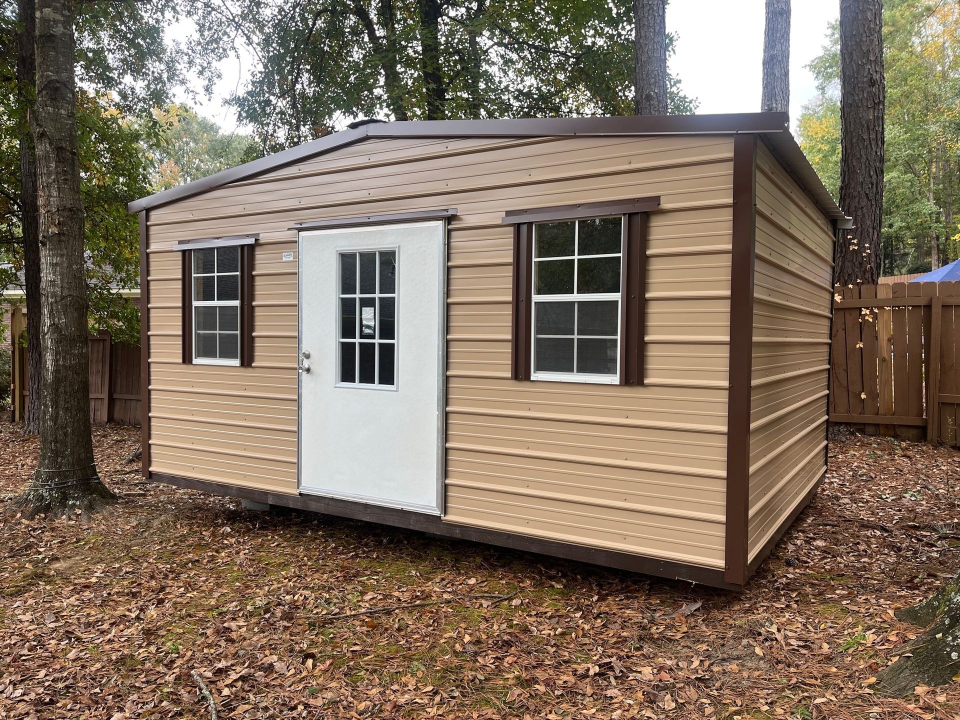 Tan metal storage shed with brown trim and roof, set in a yard with trees.