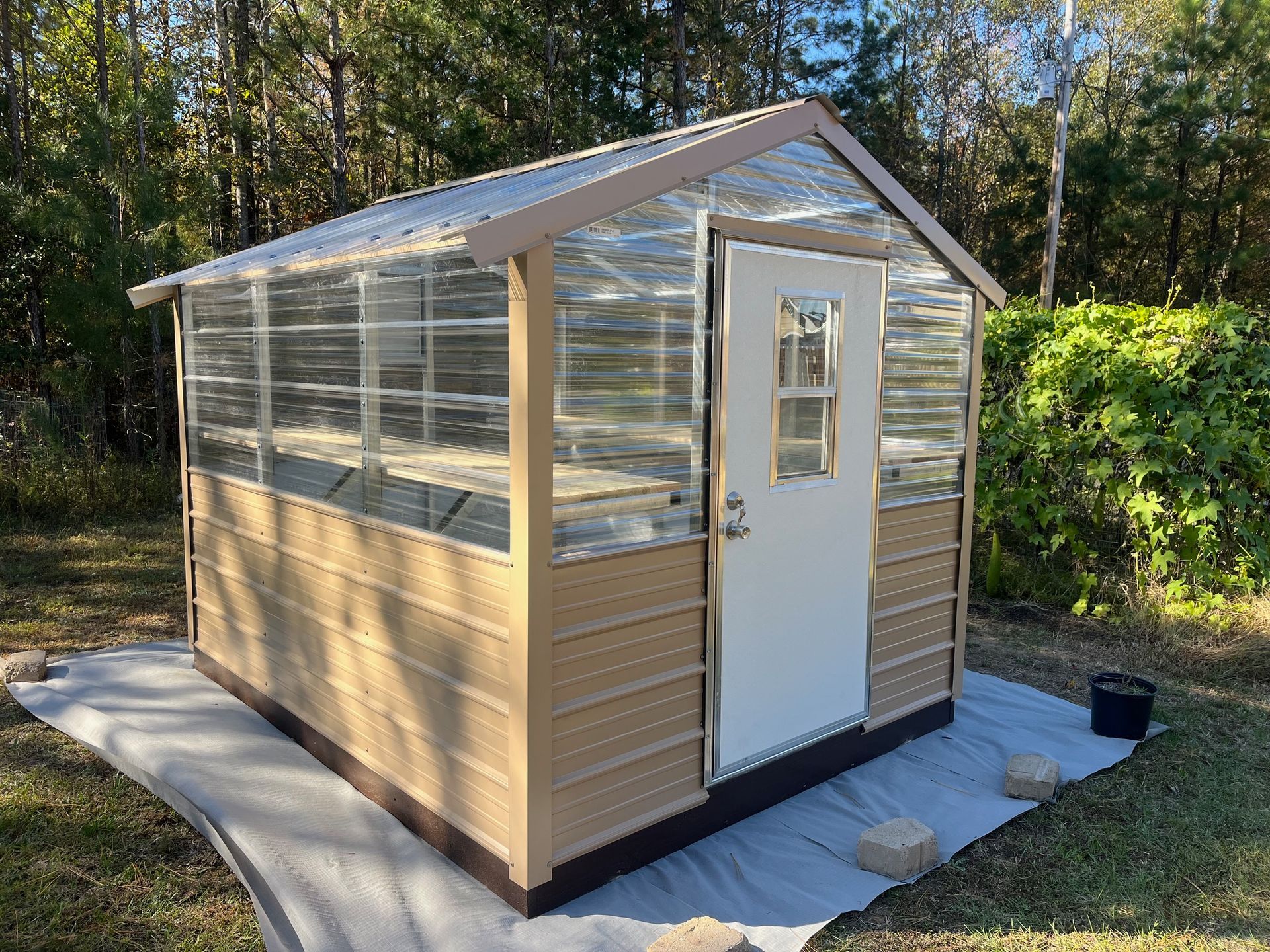 A tan and clear panel greenhouse with a white door, set on a white tarp in a grassy area.