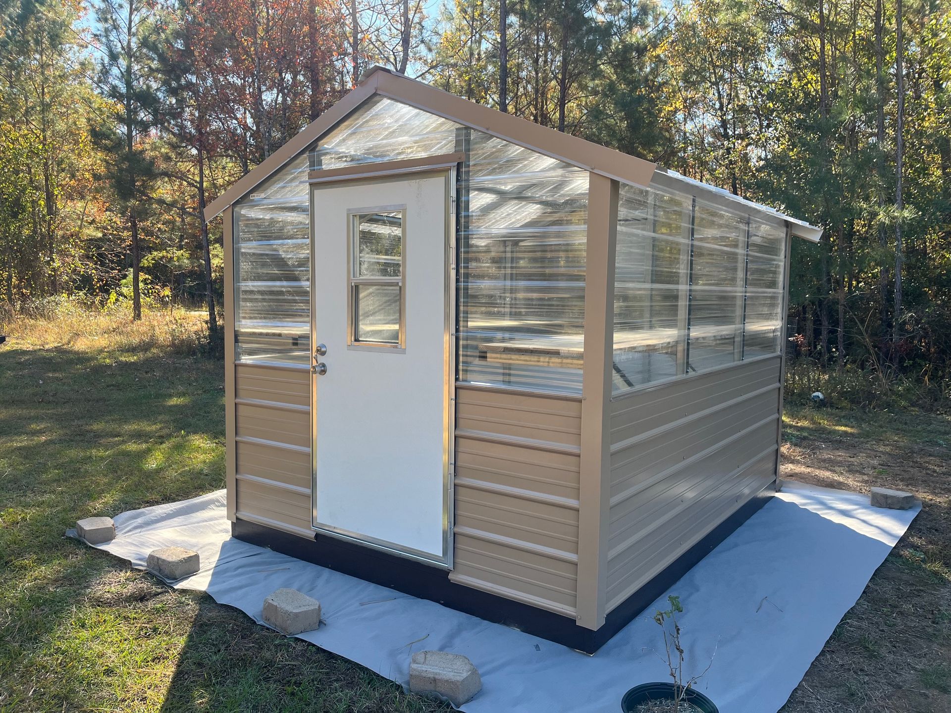 Tan greenhouse with clear panels, white door, and surrounding trees.