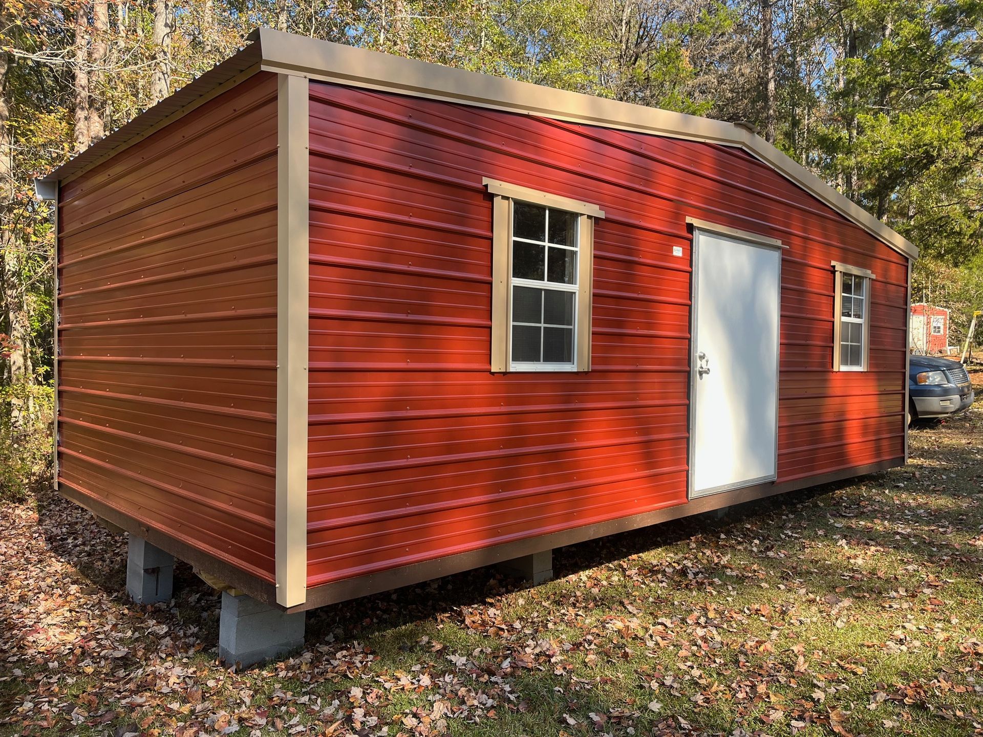 Red metal shed with a white door, two windows, and tan trim, sitting on concrete blocks, outdoors.
