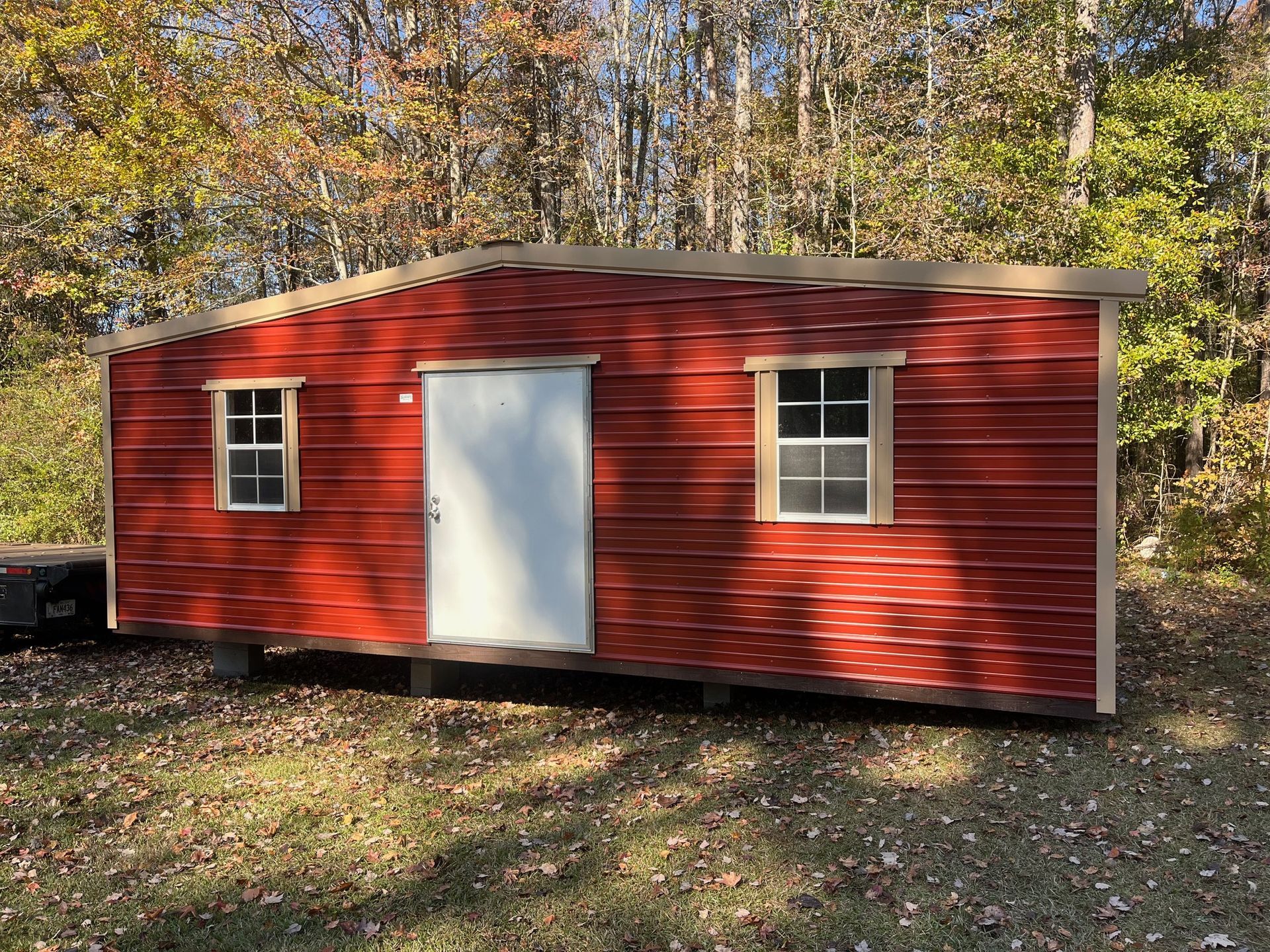 Red metal shed with white door and windows; brown trim; fall foliage in background.