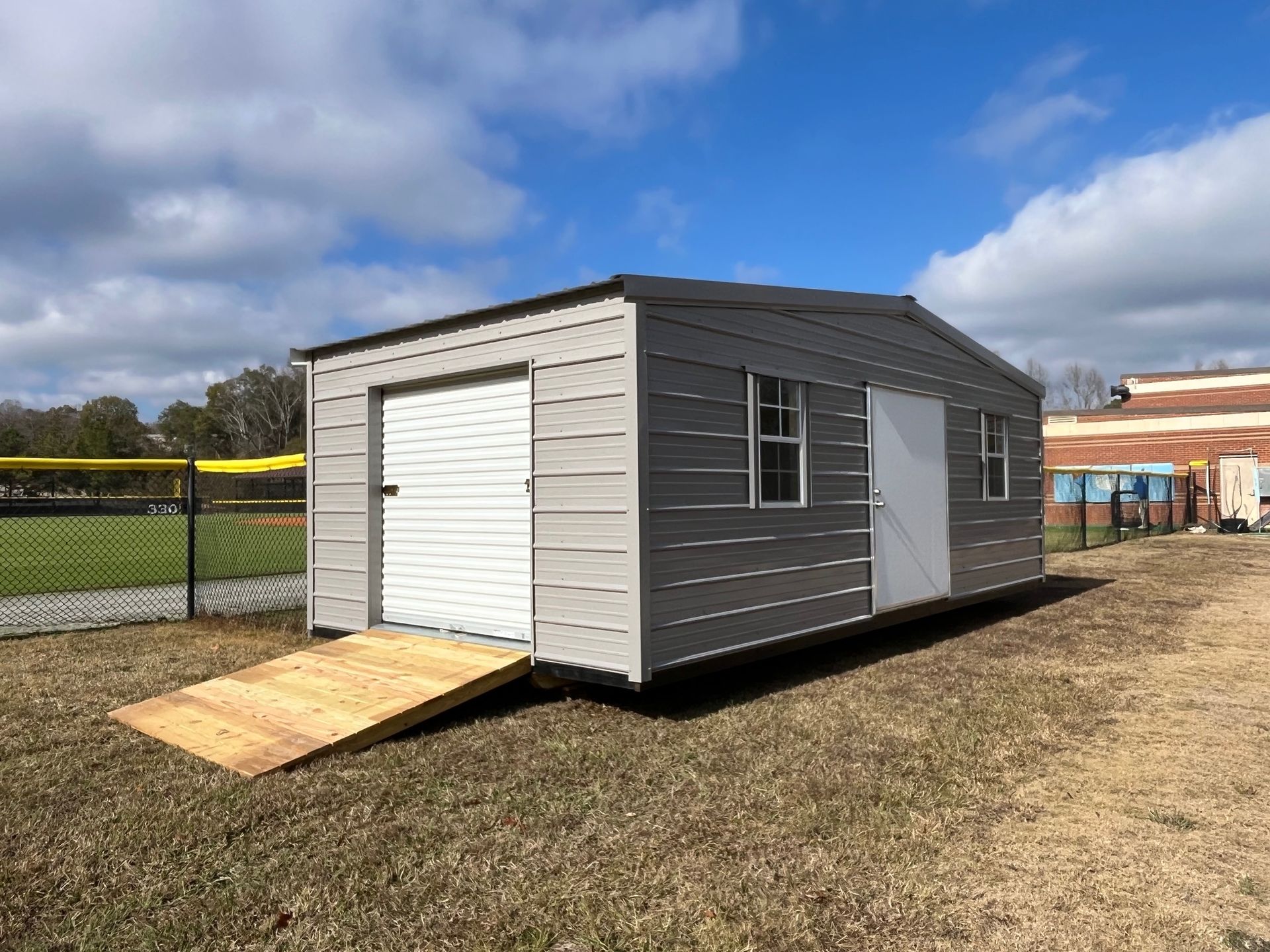 Gray metal shed with a white garage door, ramp, and side door; cloudy sky.