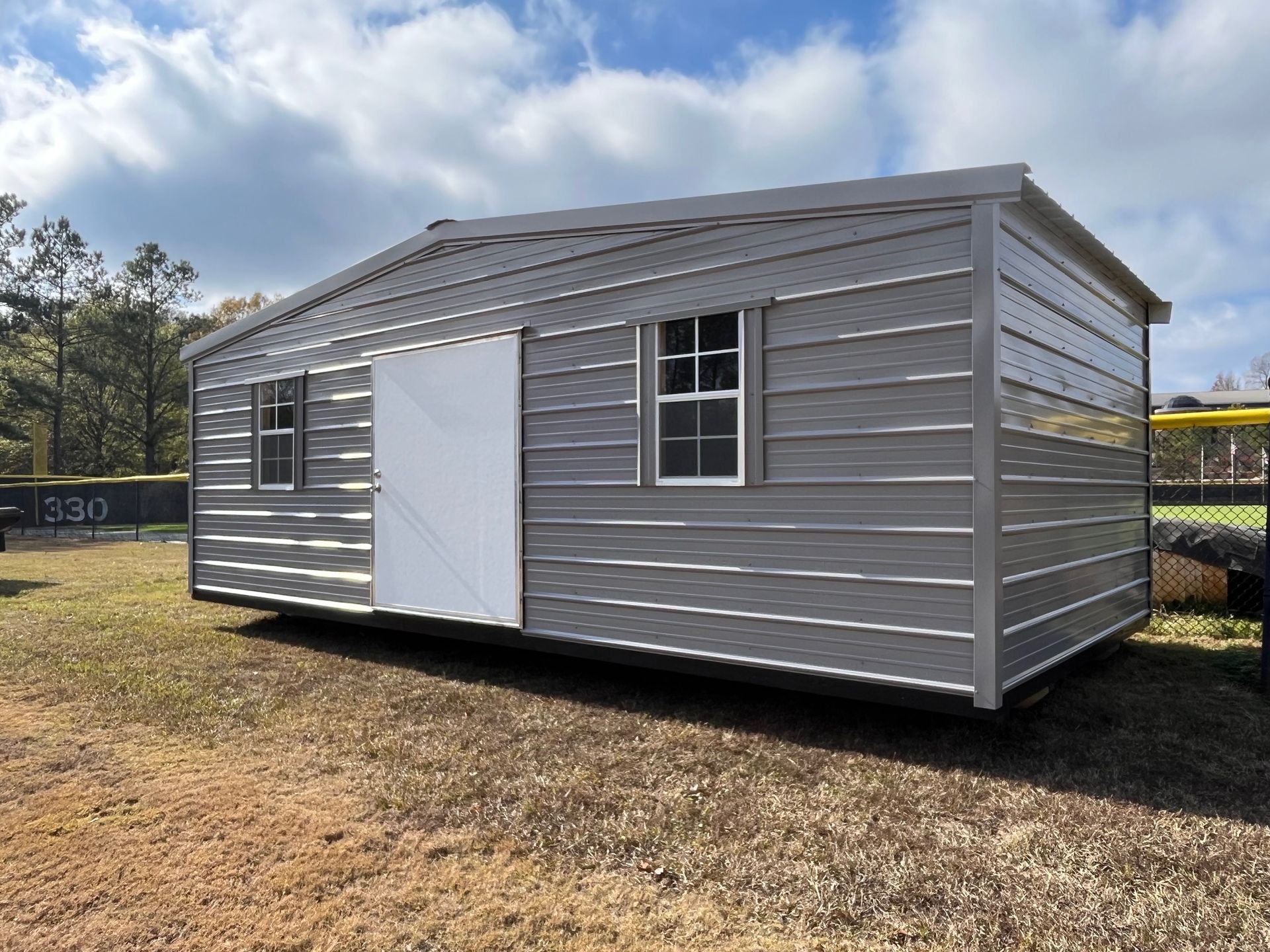 Gray metal storage shed with white door and windows, in a grassy field.