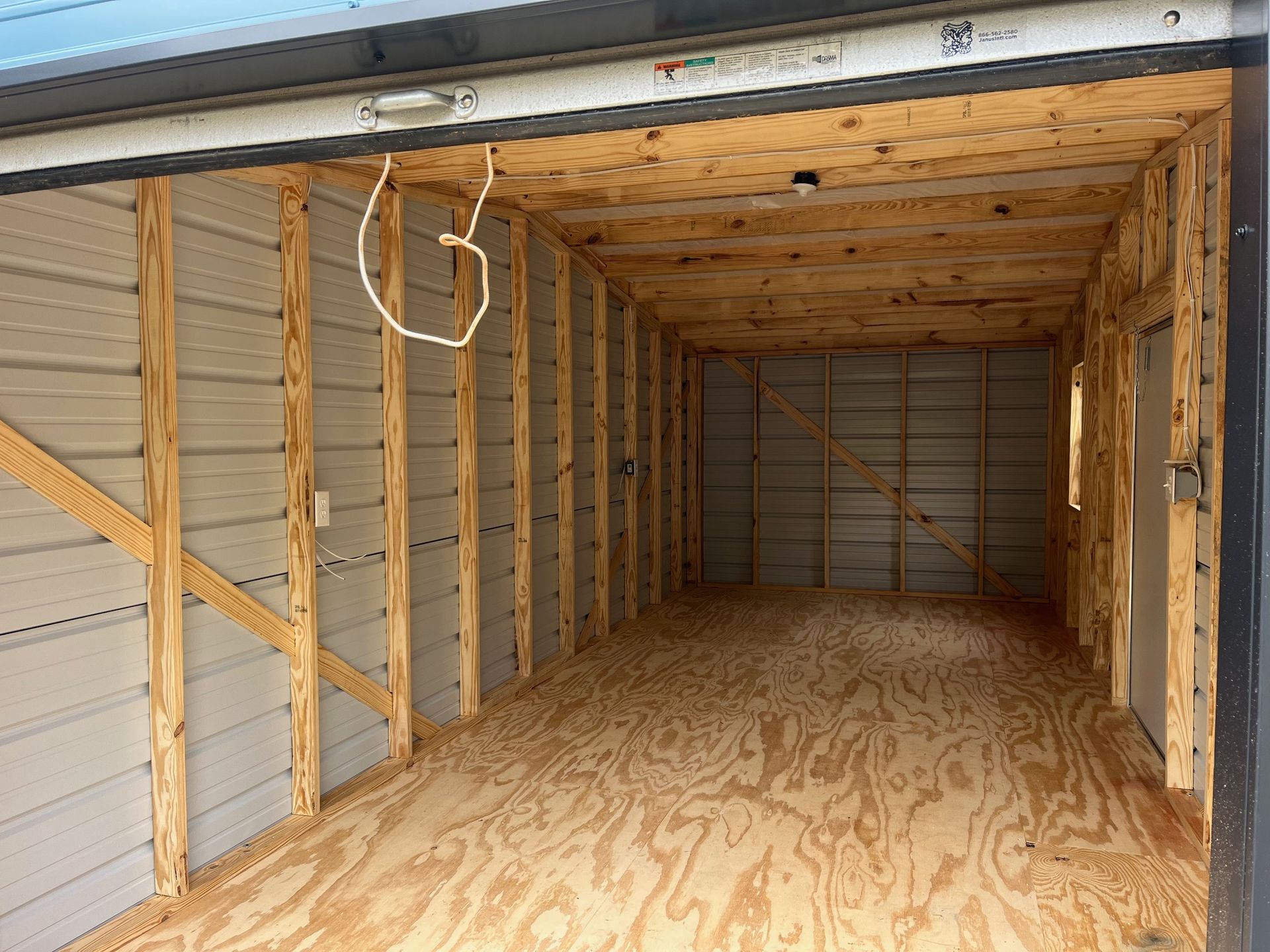 Empty garage interior with plywood floor and walls, exposed wood framing, and open garage door.