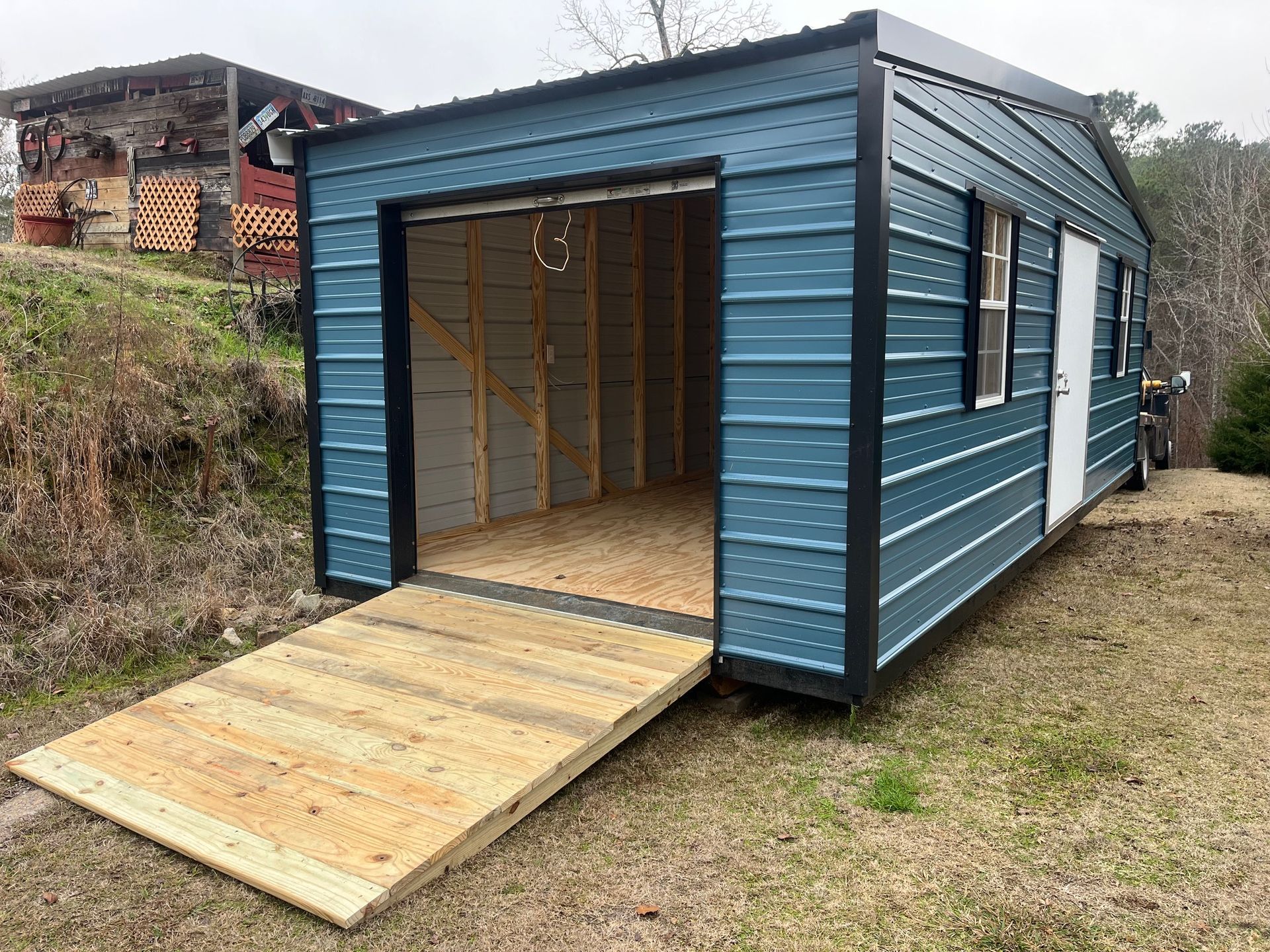 Blue shed with open garage door and wooden ramp on grass.