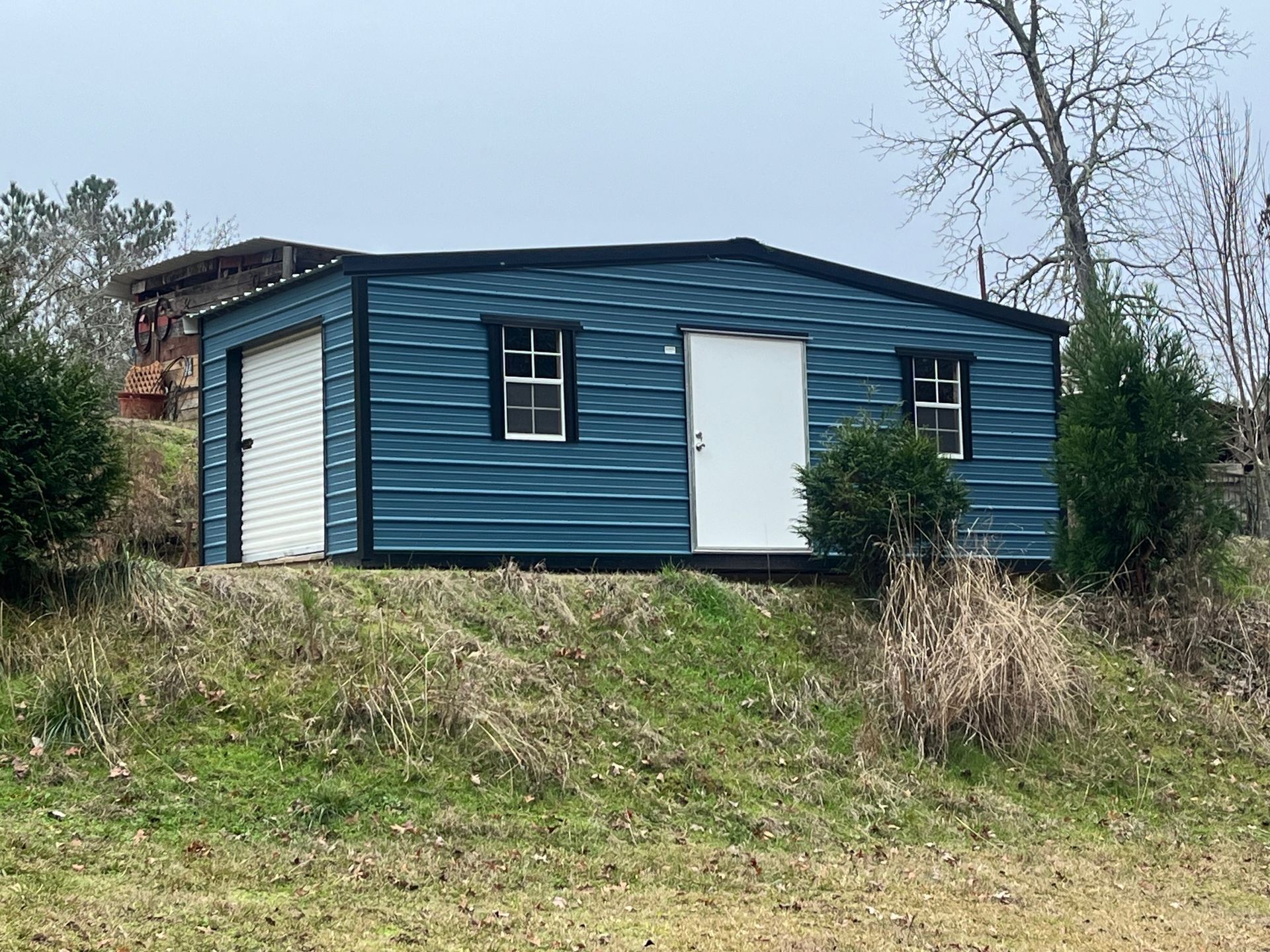 Blue metal shed with garage door, white door, and windows on a grassy hill.