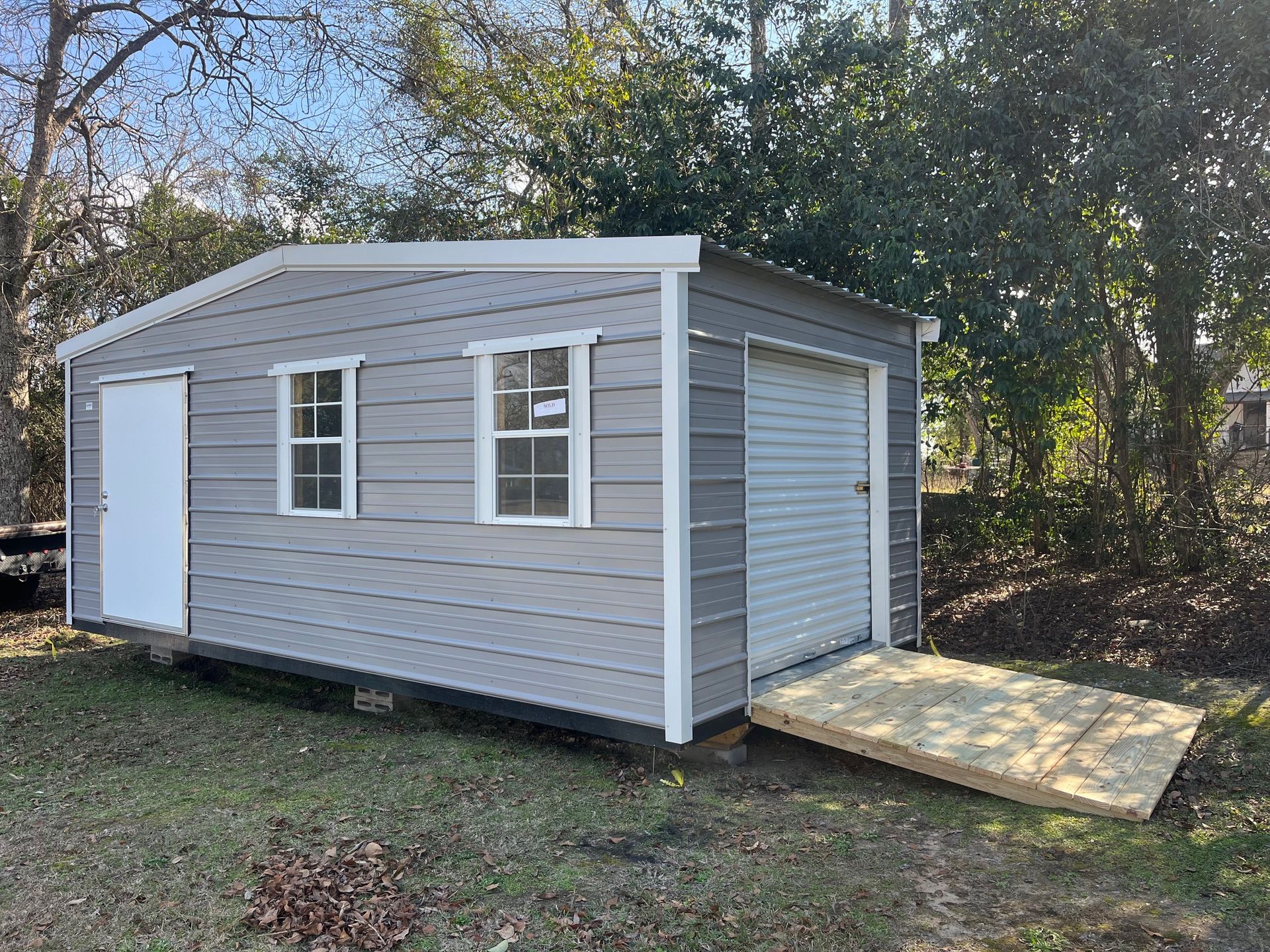 Gray shed with white trim, door, two windows, and garage door; wooden ramp in front.