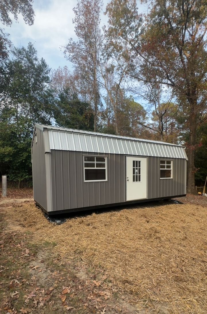 Gray shed with metal roof and white door, two windows, set in a yard with dry leaves.