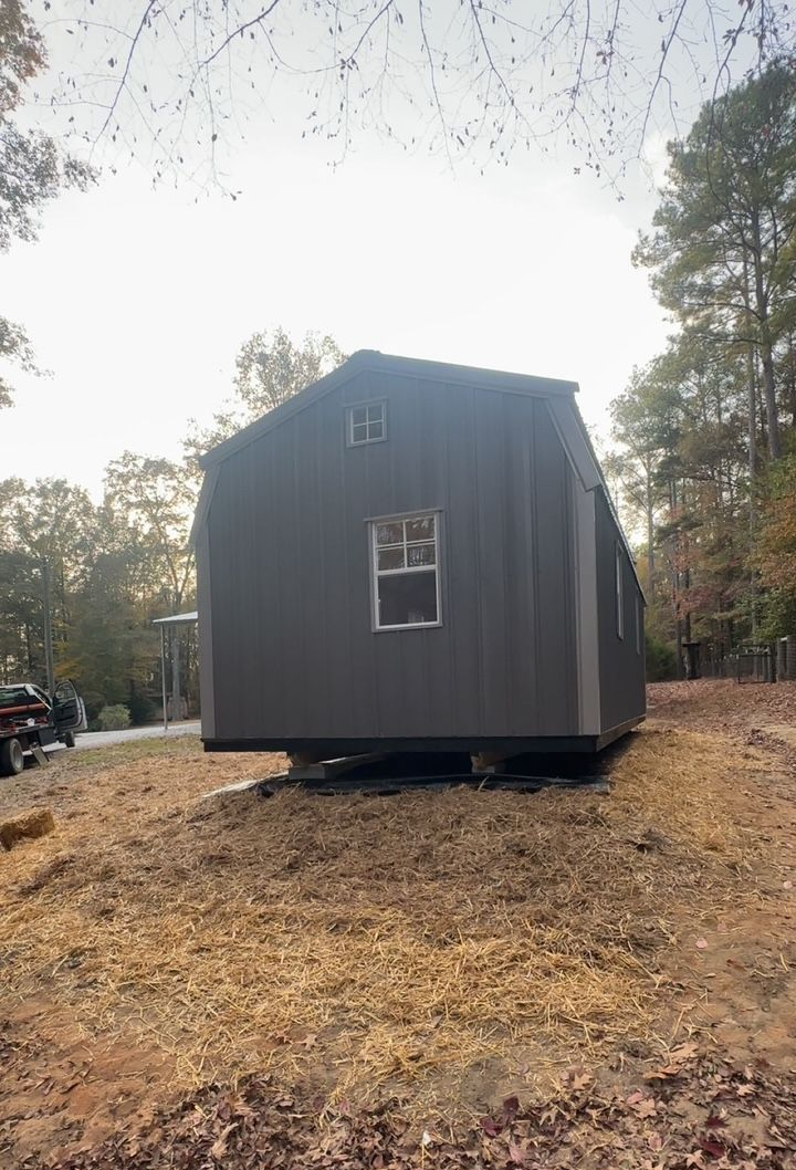 Dark gray tiny house on a dirt lot, framed by trees under an overcast sky.