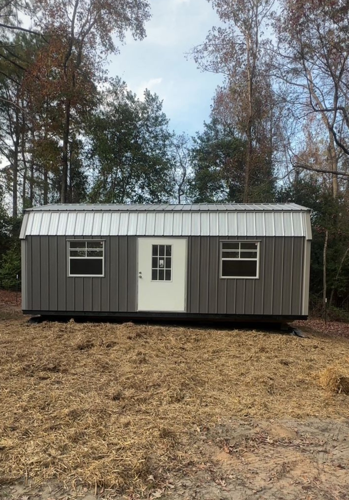 Gray metal storage shed with white door and windows, sits in a clearing.