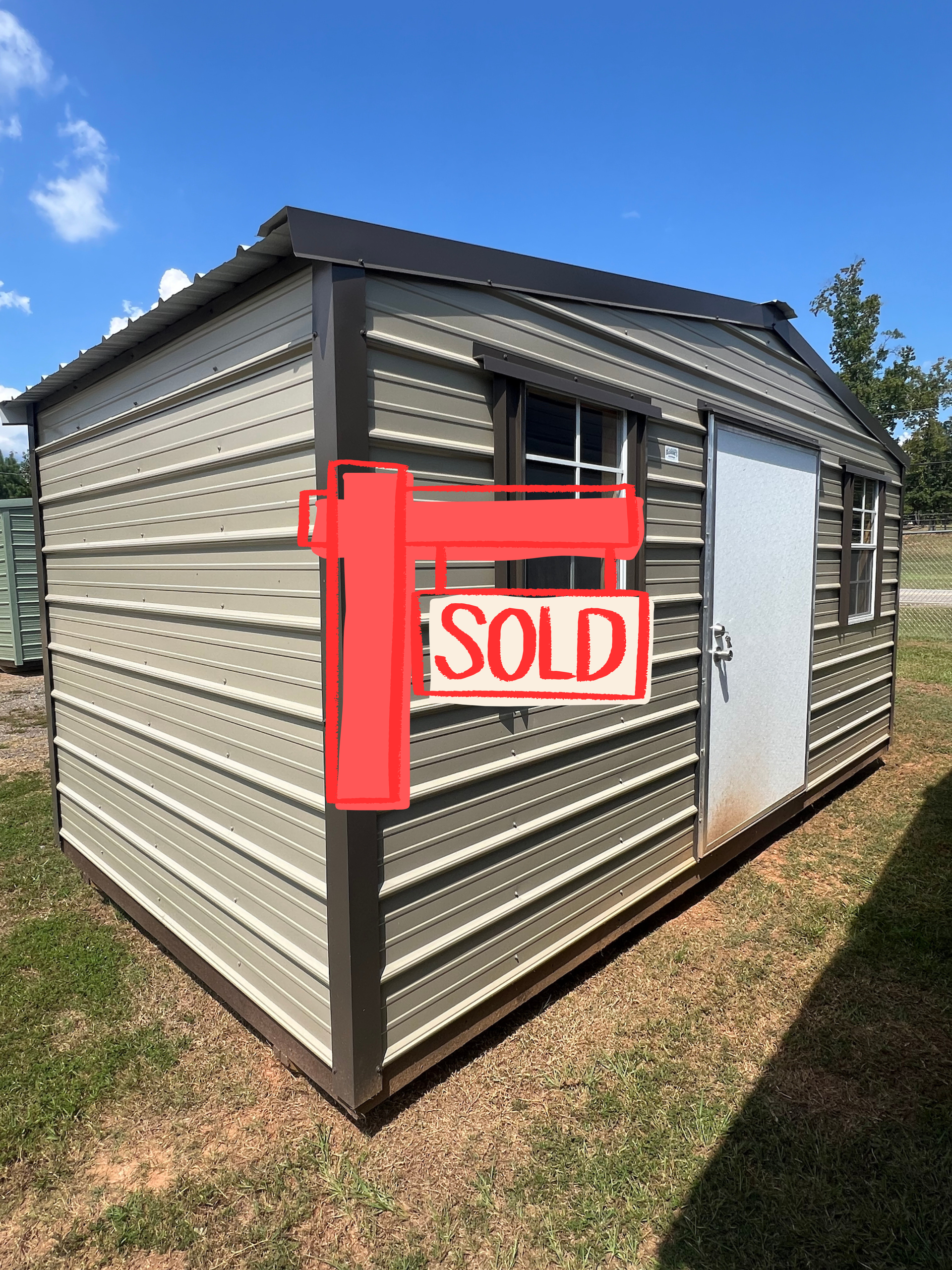 Tan and brown metal shed with a black roof, two windows, and a white door, on grassy ground.