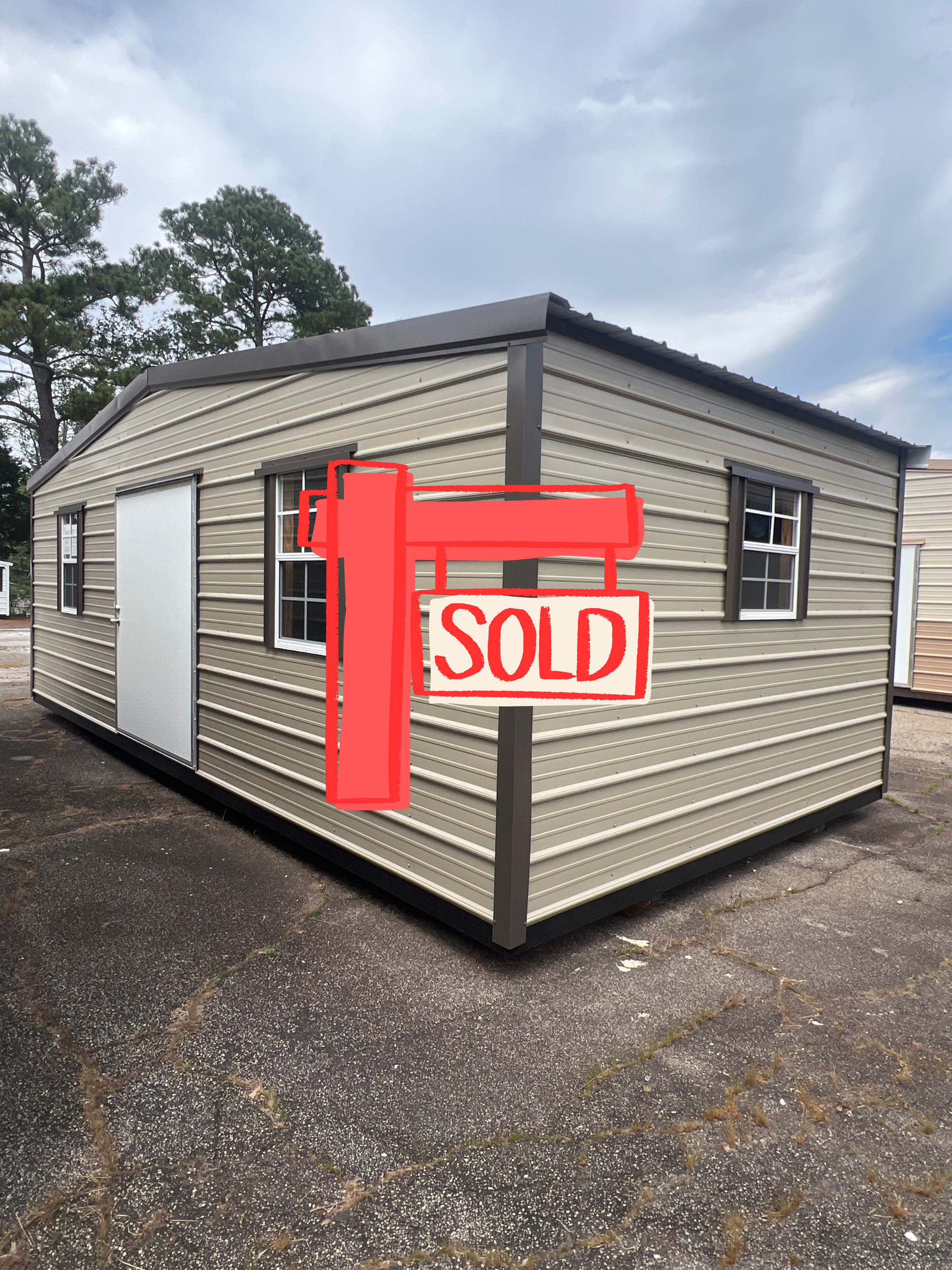 Small beige mobile home with a red “SOLD” sign outside on a gravel lot under cloudy skies