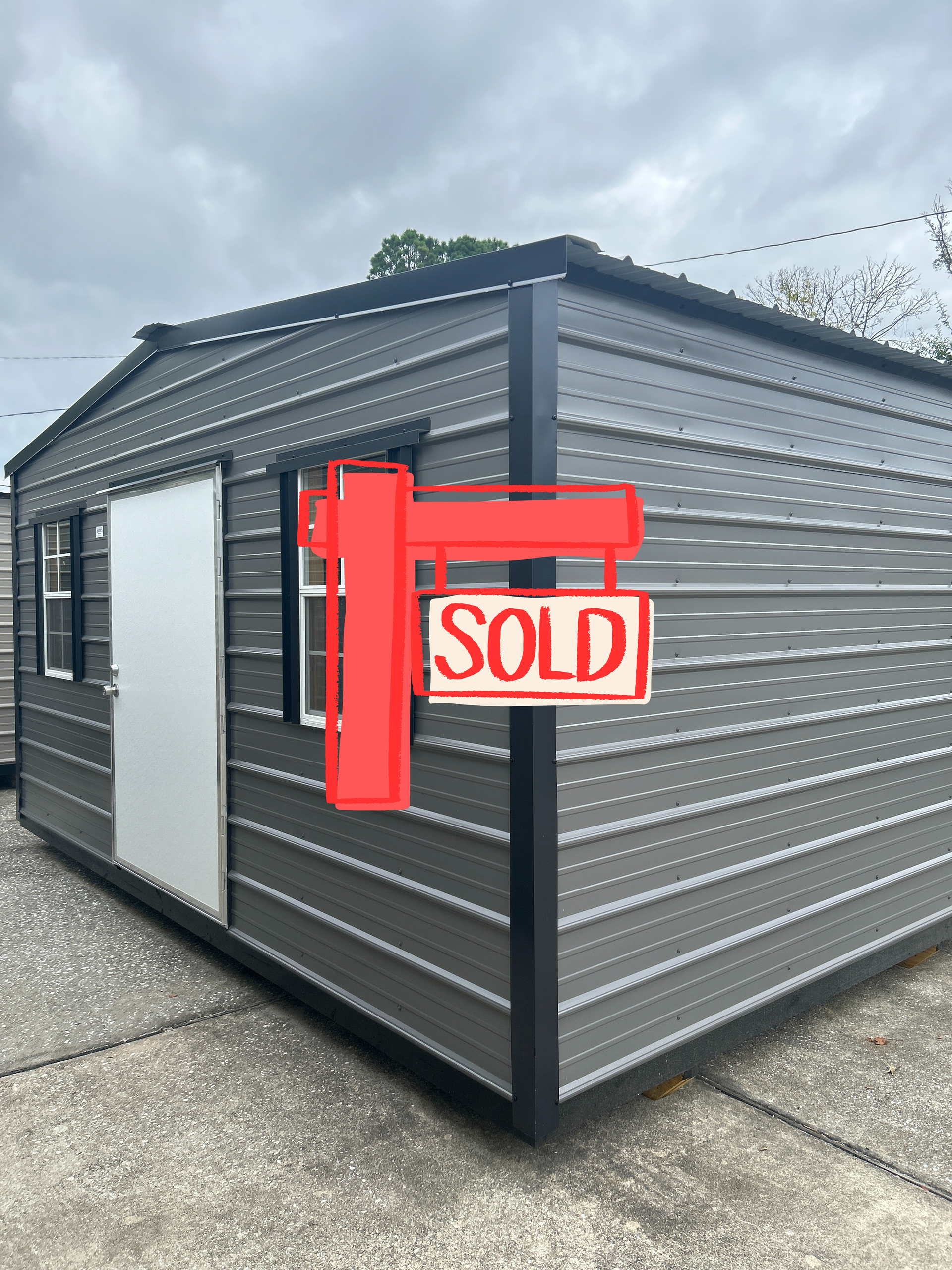 Dark gray metal shed with a white door, two windows, and black trim, outdoors.