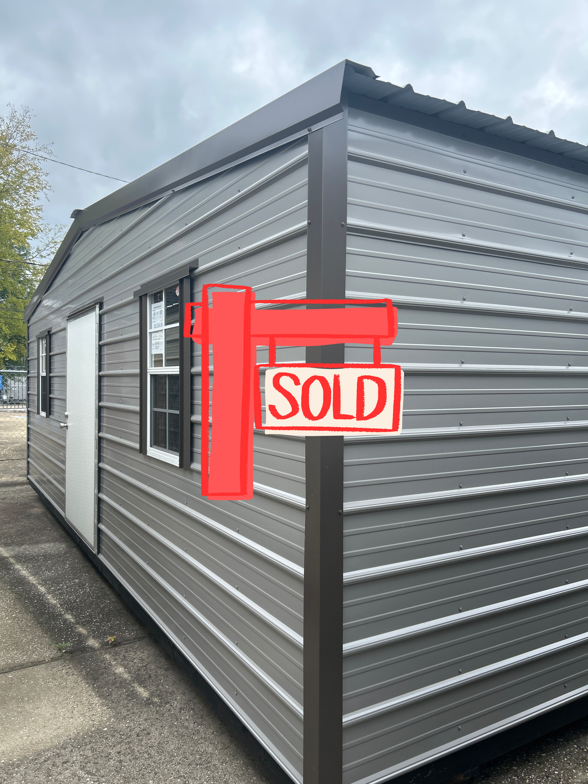 Gray metal shed with door and windows, on a paved surface, overcast sky.