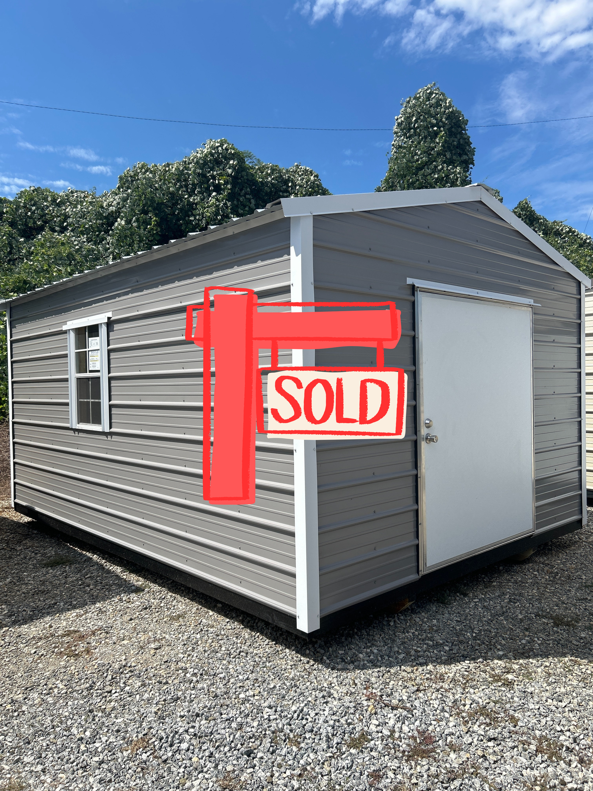Gray metal storage shed with a white door and window, set on gravel.