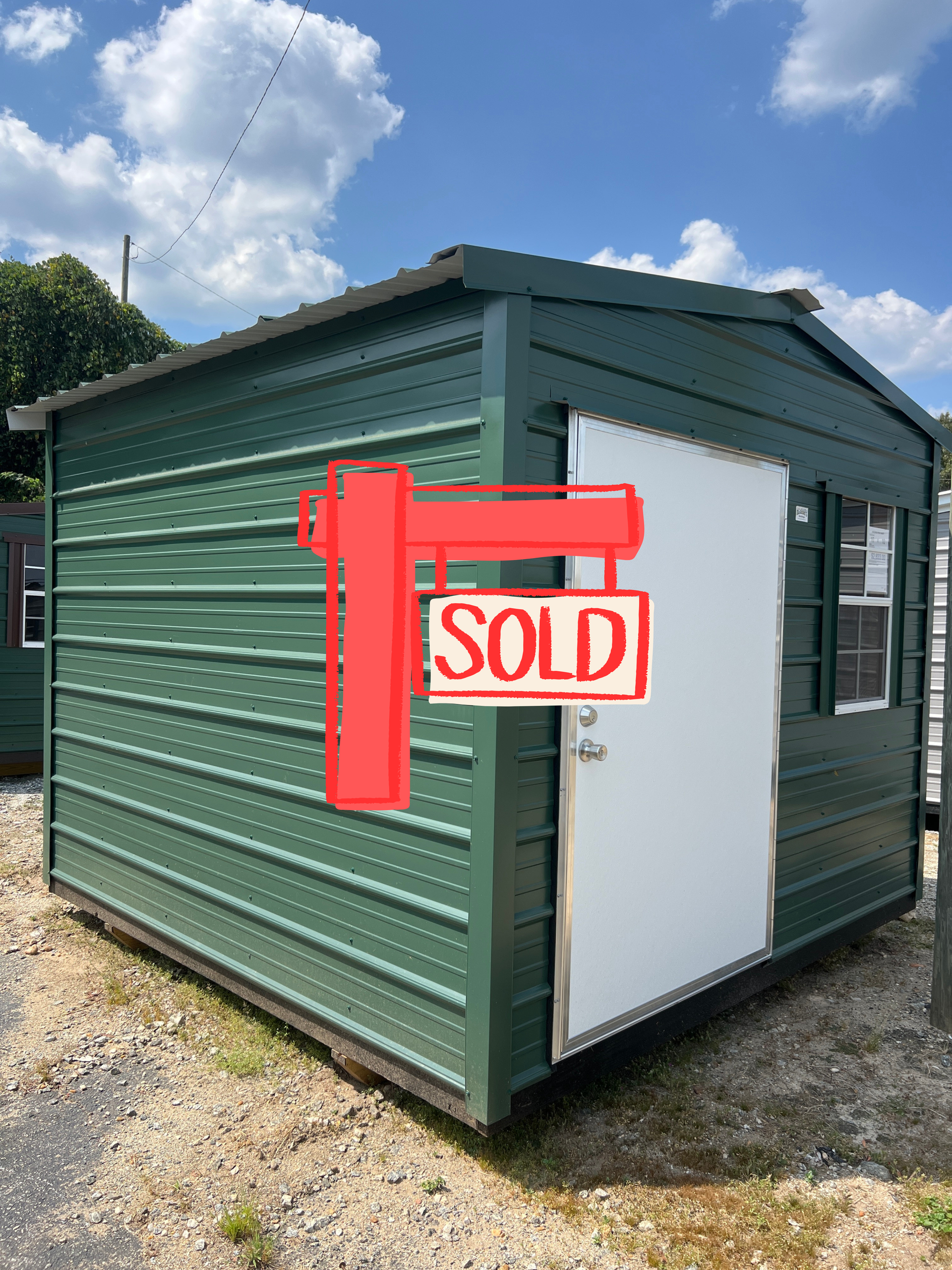Green metal shed with white door and window, sitting outside on a gravel lot.