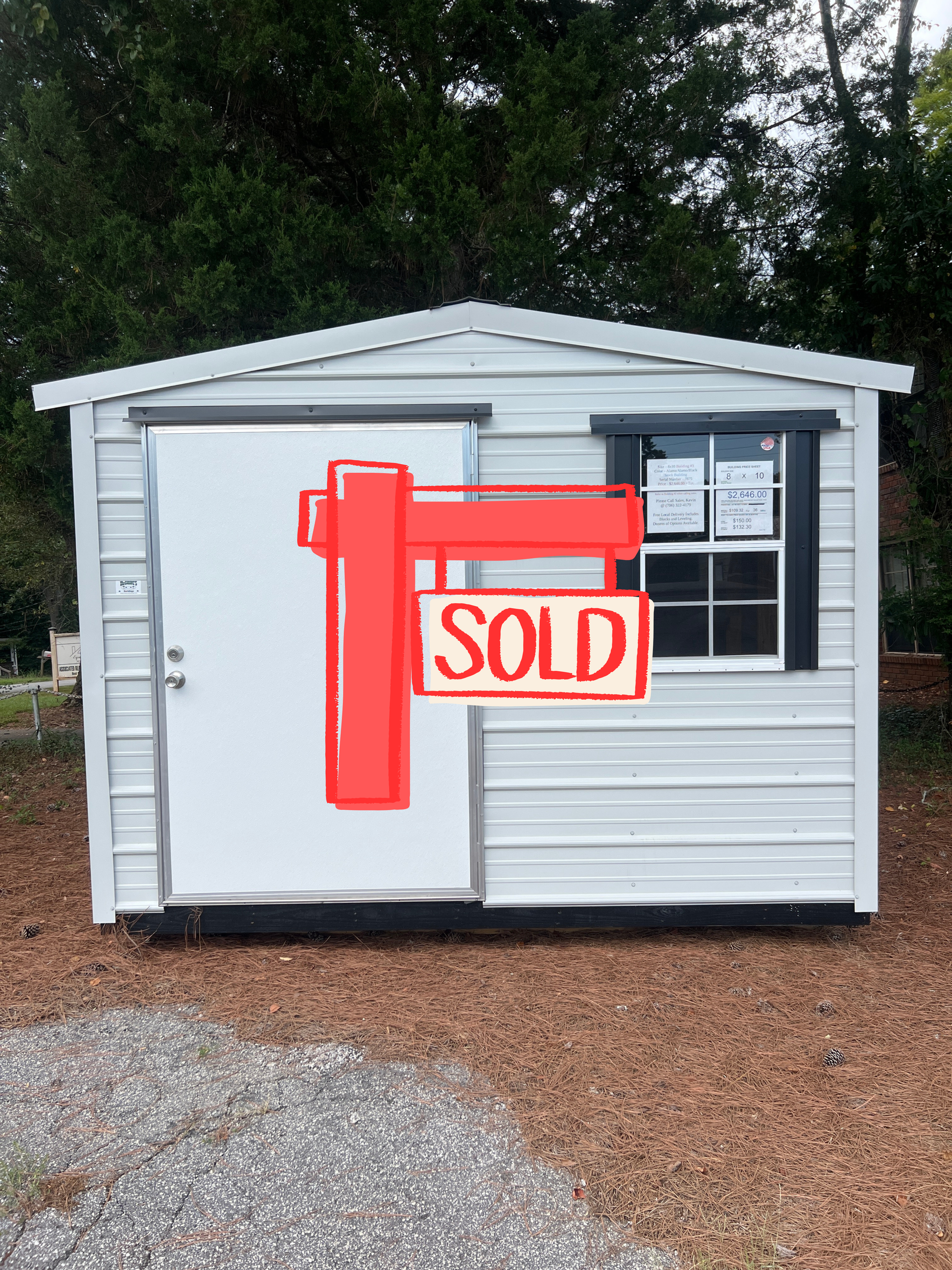 White shed with a sliding door, a black-framed window, on a gravel and pine straw surface.