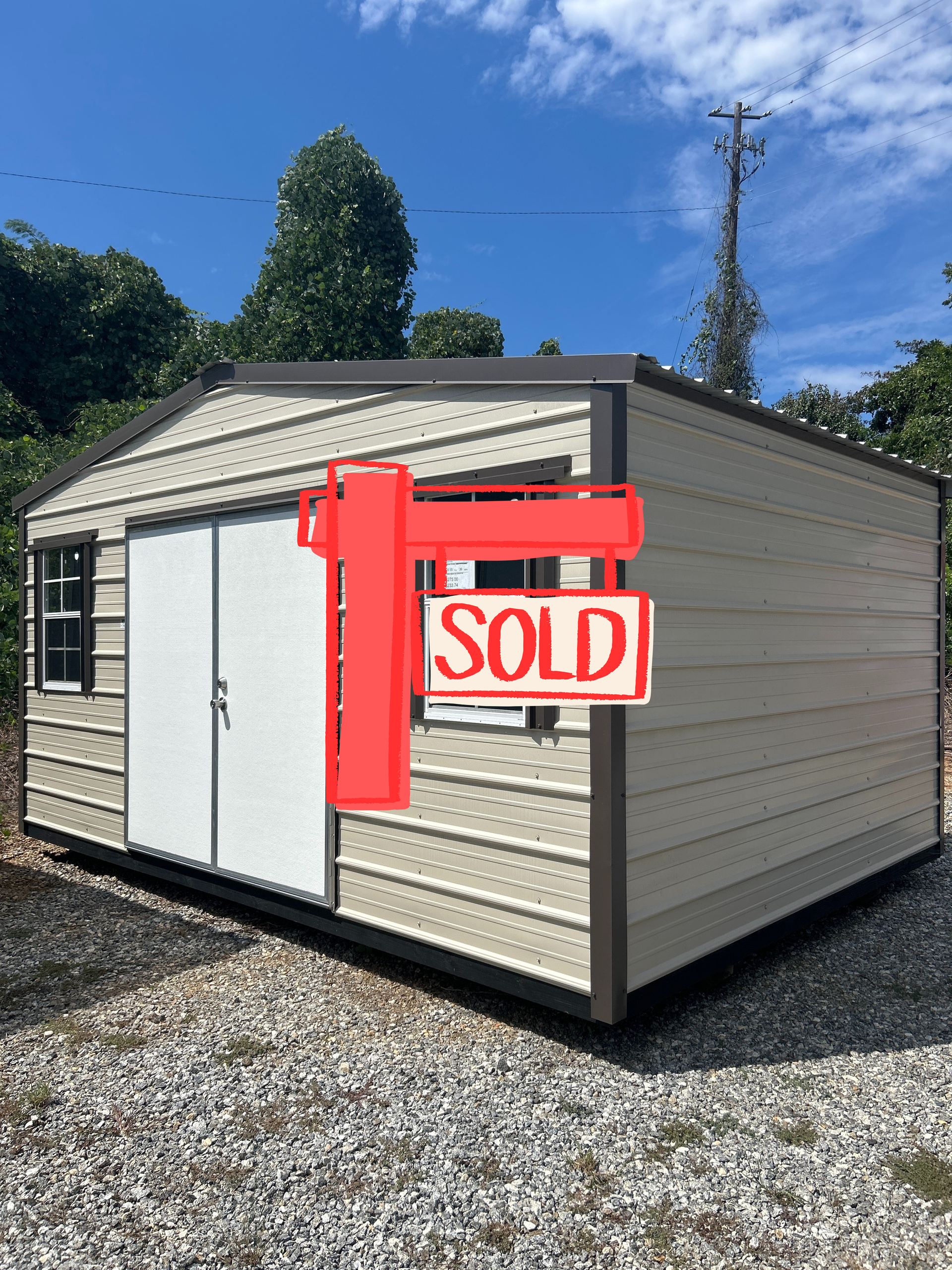 Tan metal storage shed with brown trim, double doors, two windows, gravel ground, and trees.
