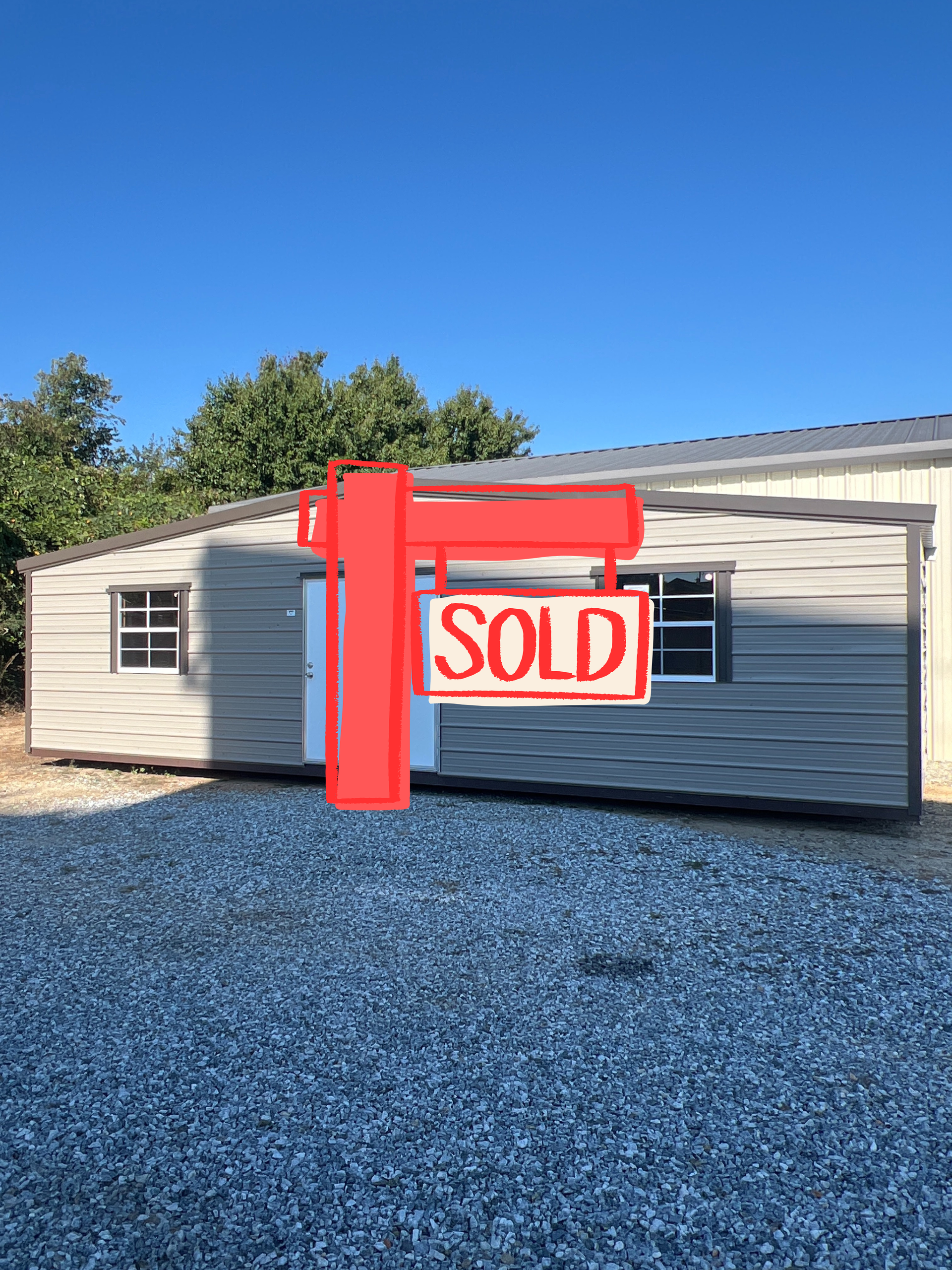 Gray shed with white door and two windows on a gravel surface under a blue sky.