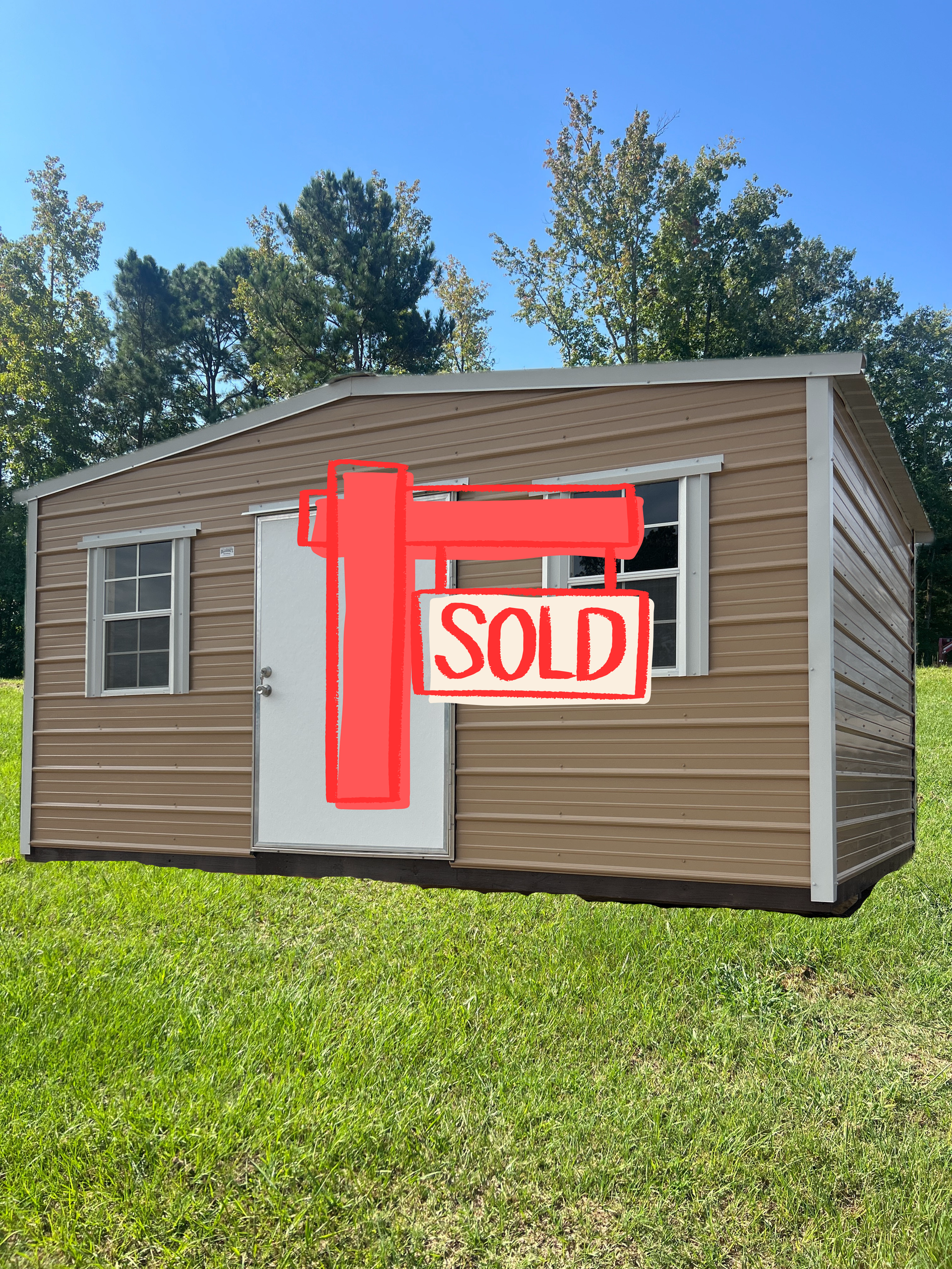 A tan, metal-sided storage shed with a white door and two windows sitting on green grass against a backdrop of trees.