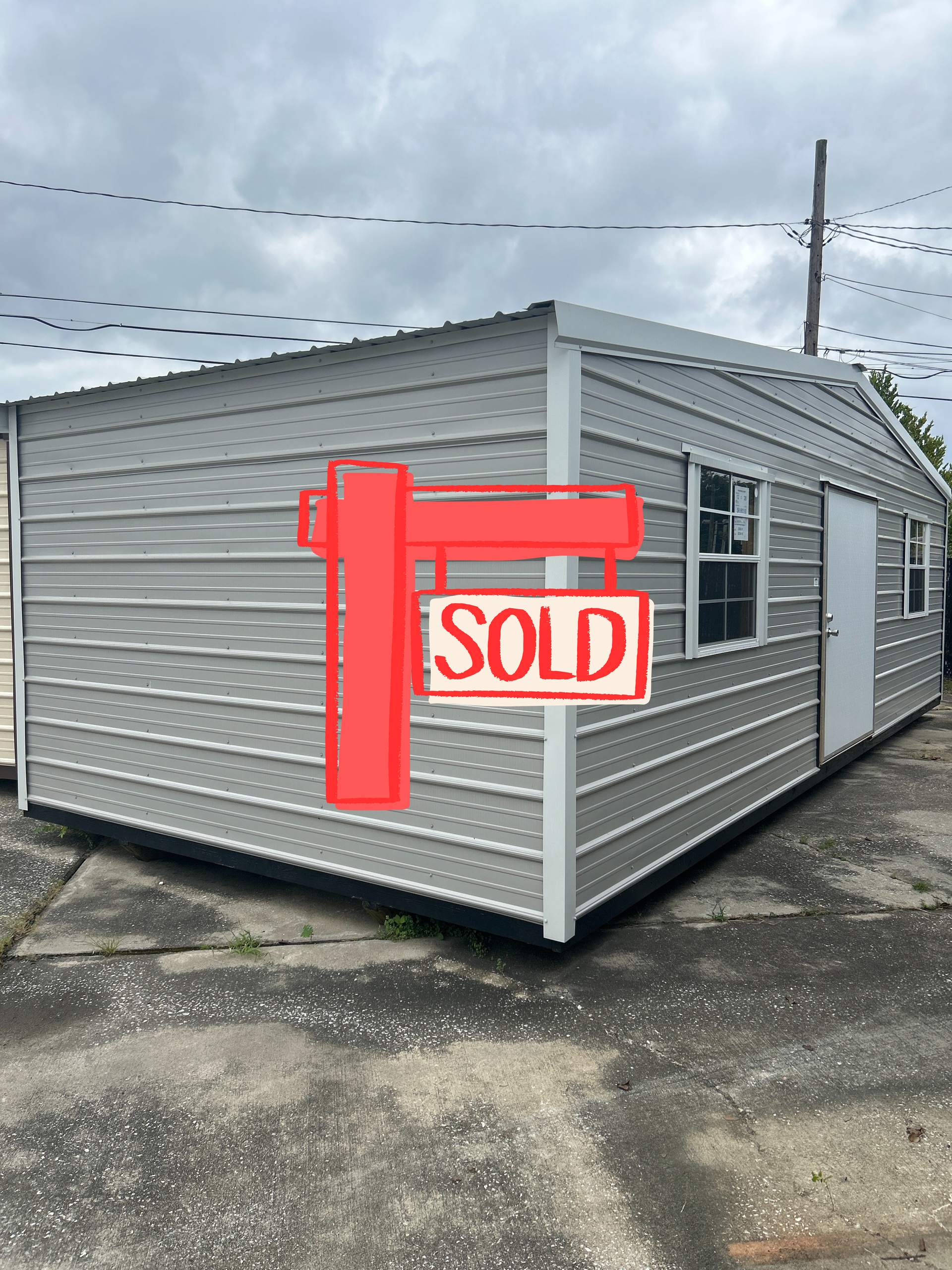 Gray metal storage shed with a white door and two windows, under an overcast sky.