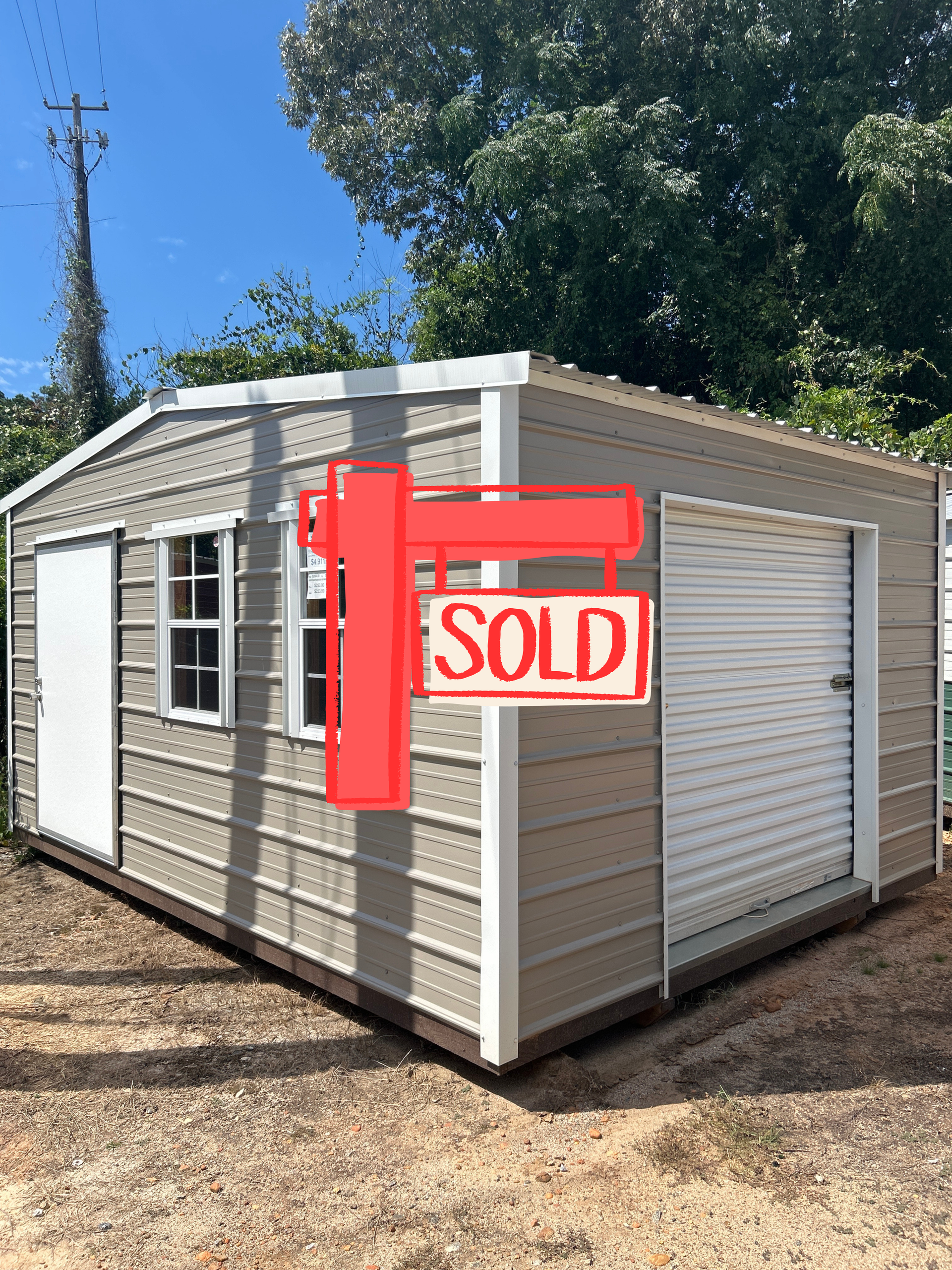 Small beige storage shed with a red SOLD sign on a gravel lot under a blue sky