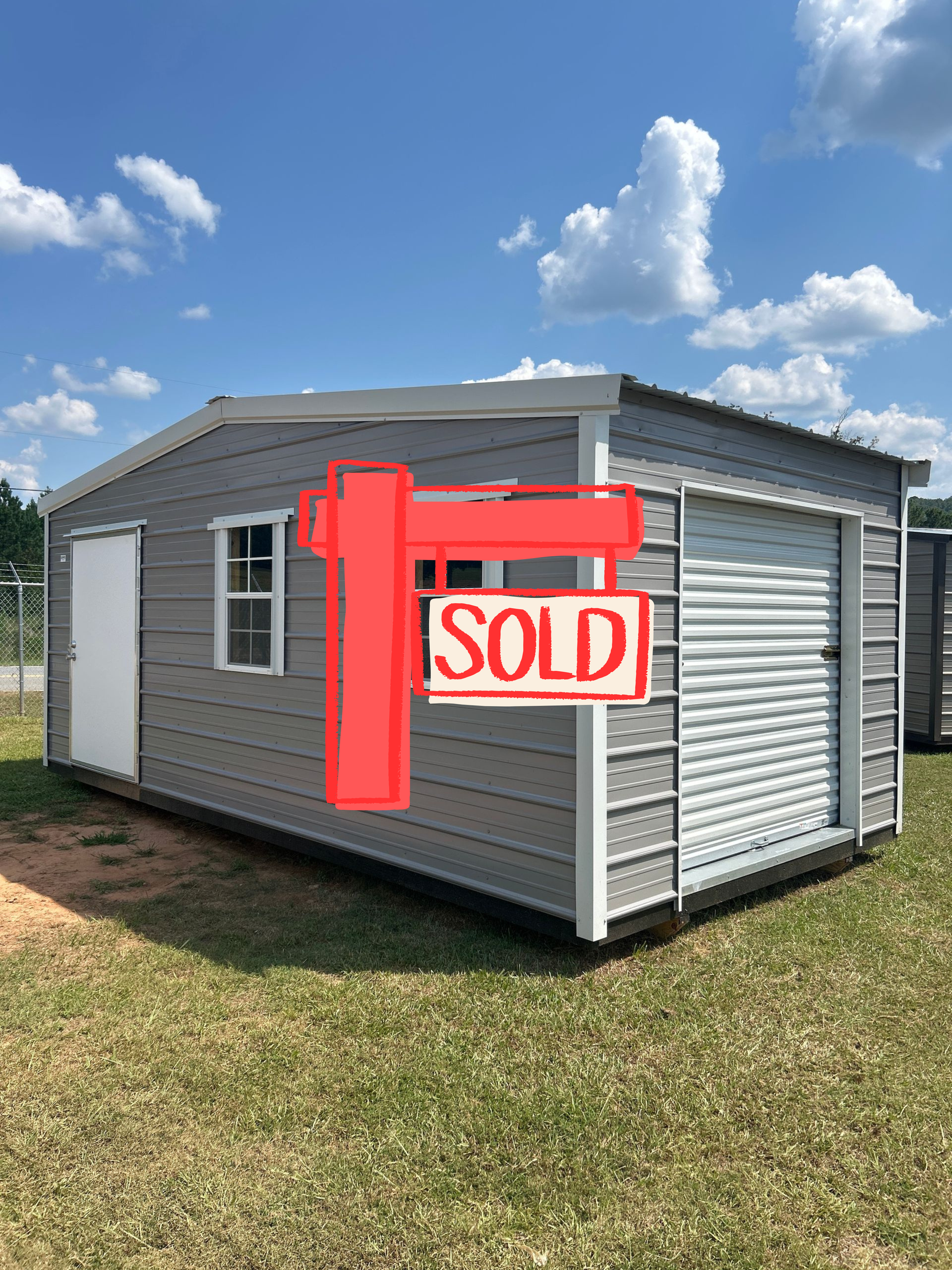 Gray shed with white trim, door, windows, and roll-up garage door on a grassy area under a blue sky.