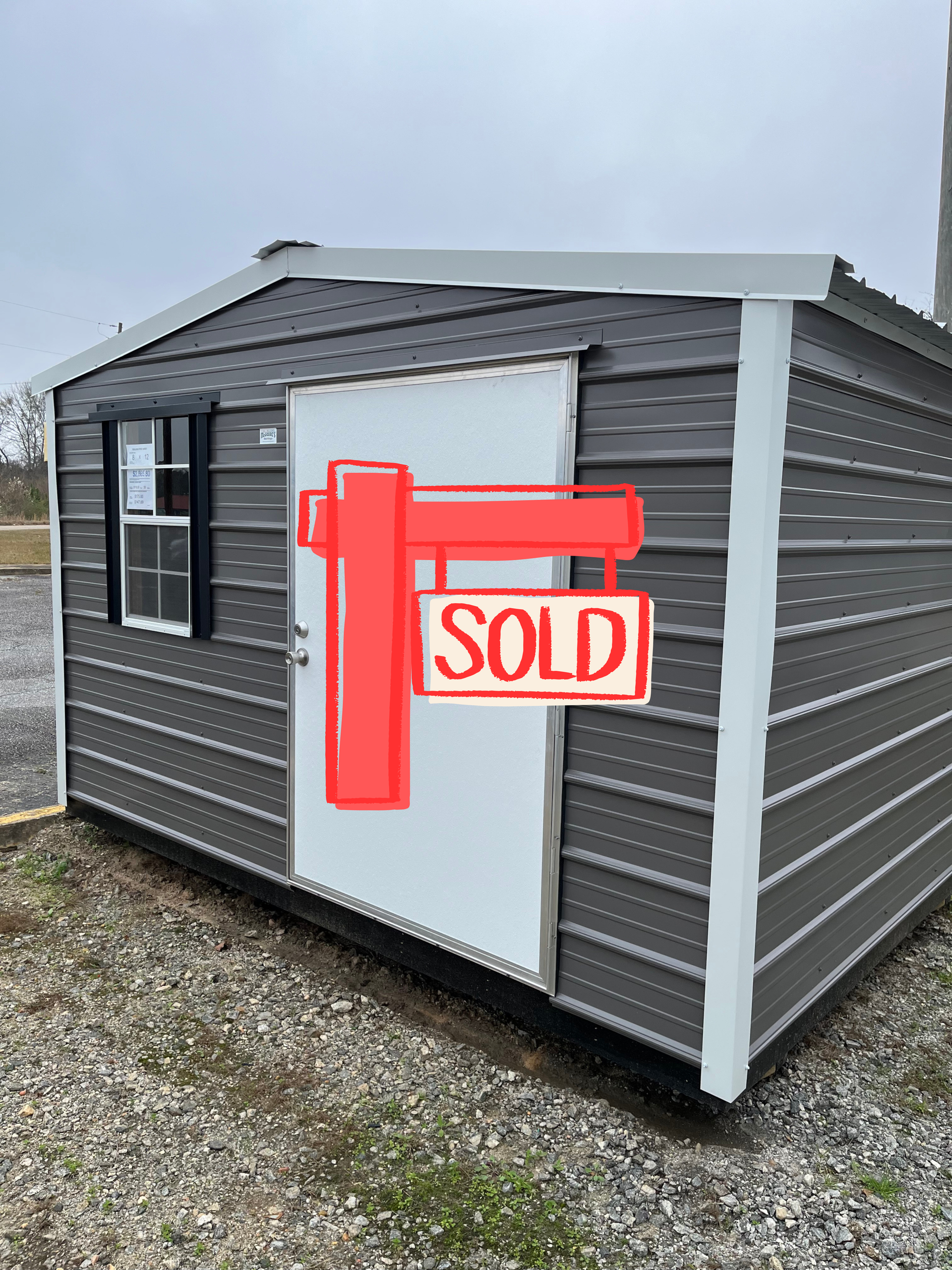 Gray metal shed with a white door and window, set on gravel.