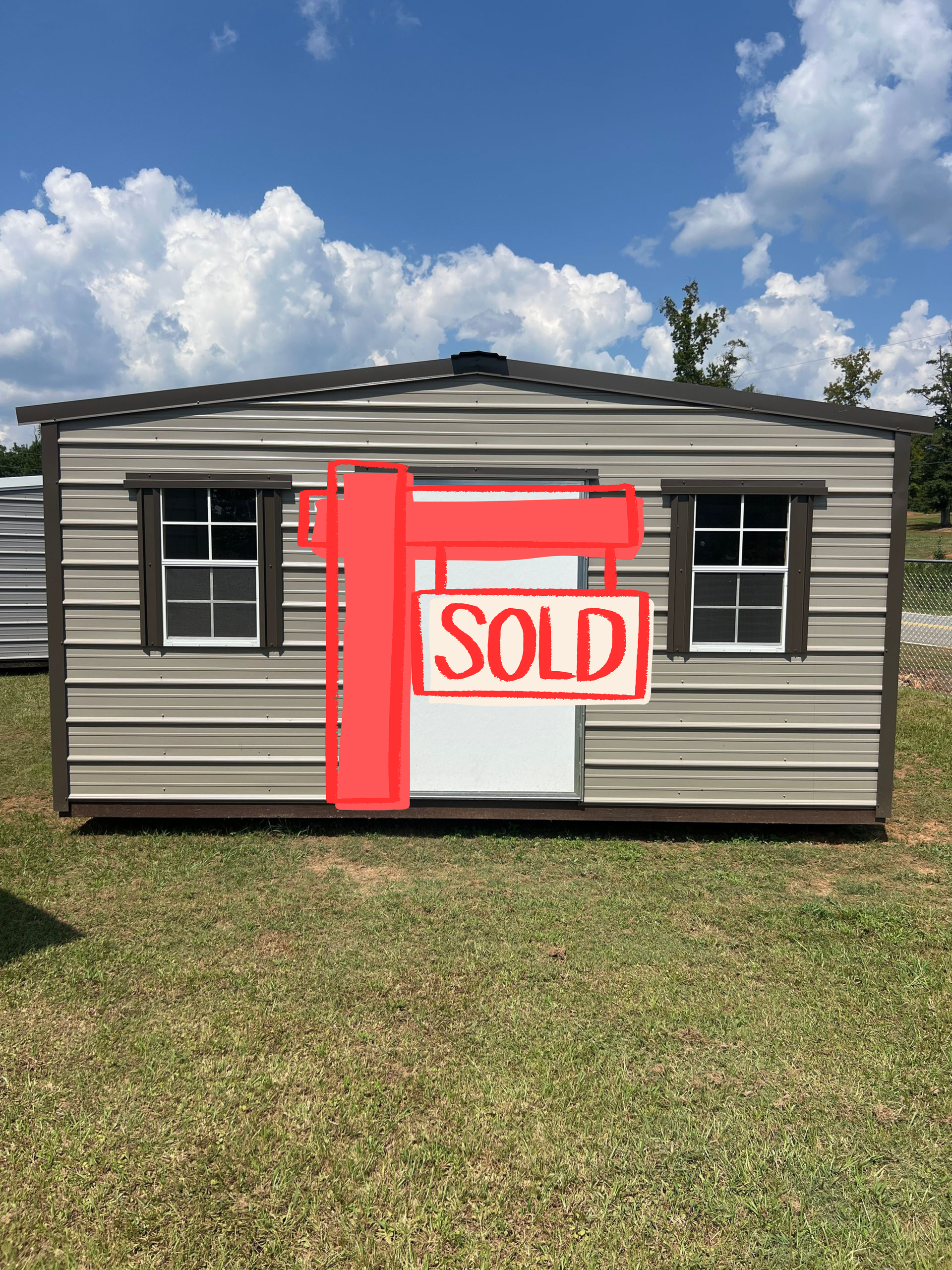 Tan metal storage shed with a white door and two windows, set outdoors on grass under a blue sky.