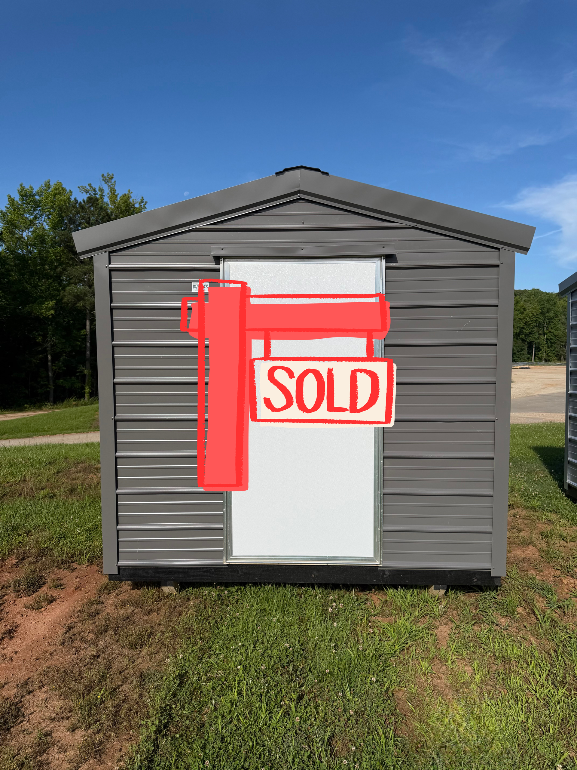 Small gray shed with a white door and red SOLD sign in a grassy yard under a blue sky