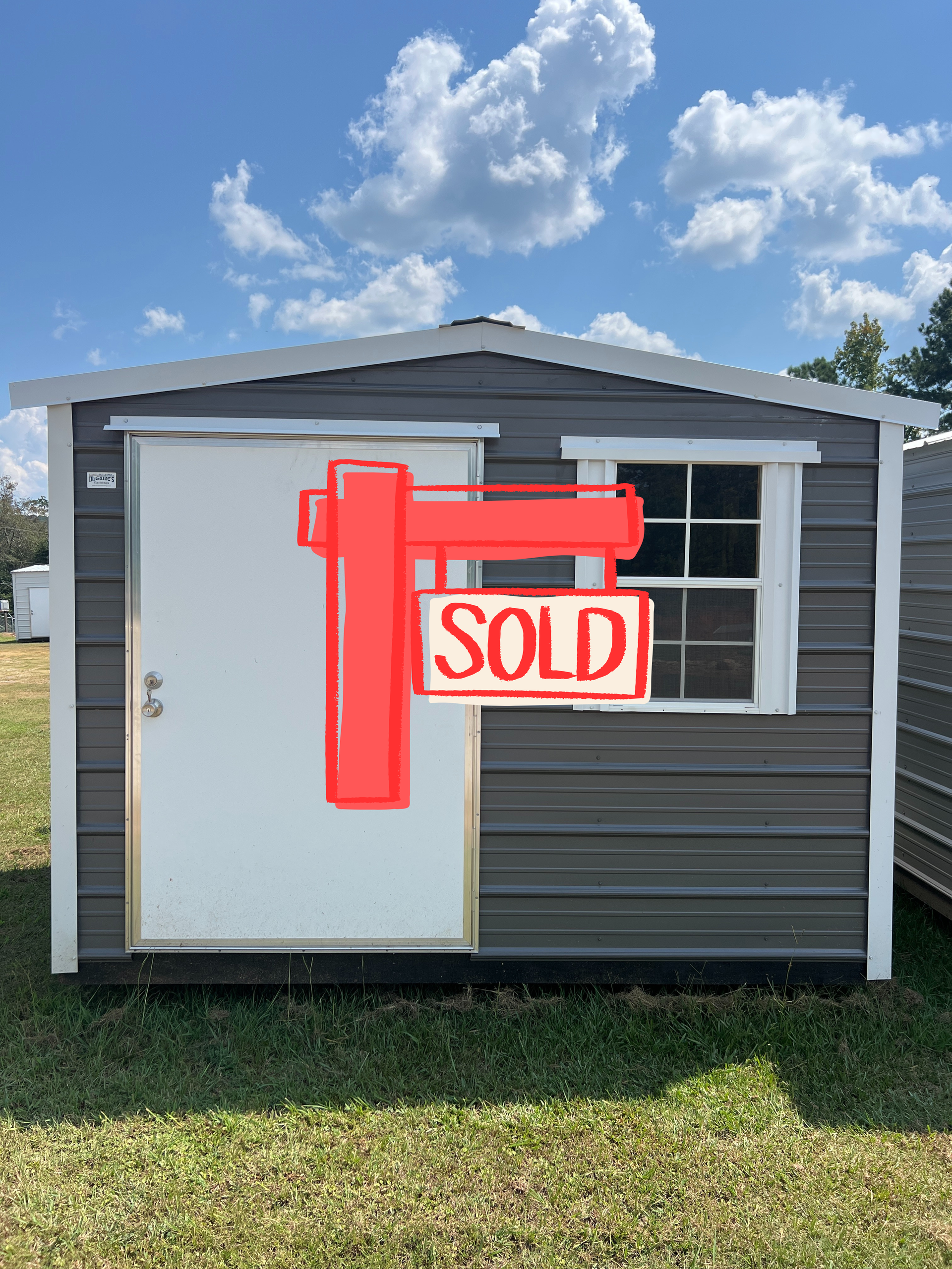 Gray metal shed with white door, window, and trim, on grass under a blue sky with clouds.